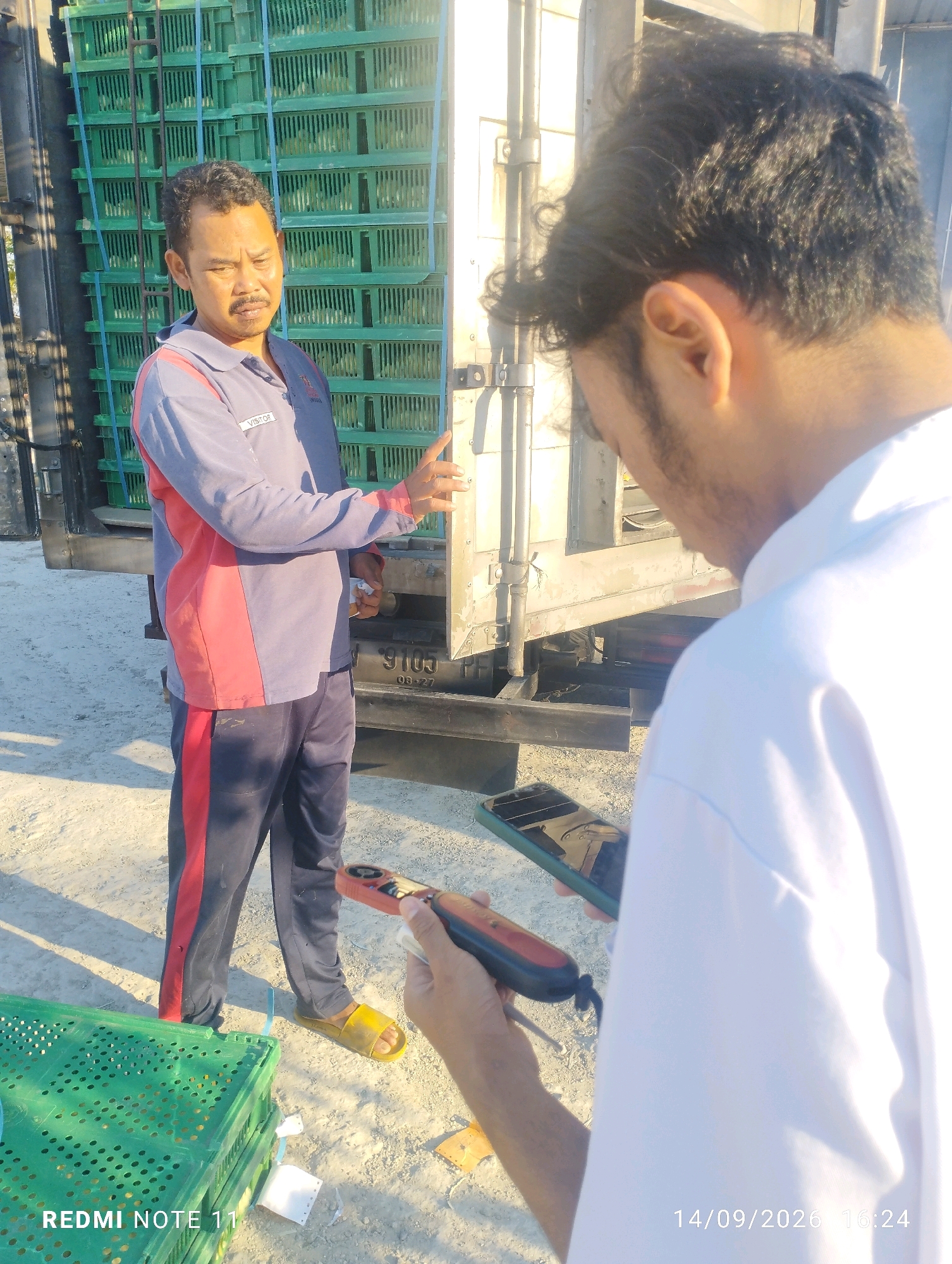 In Bero, Indonesia, a man wearing a blue and red uniform with a visitor's tag, dark pants, and yellow sandals stands in front of a truck filled with green crates. He is gesturing with his right hand towards the truck. Another person, seen from the back and wearing a white shirt, is holding a handheld device, possibly a scanner, and a smartphone. The ground is covered with light-colored sand, and sunlight casts shadows. A green plastic crate is visible in the foreground on the left. The image is dated 14/09/2026 at 16:24.