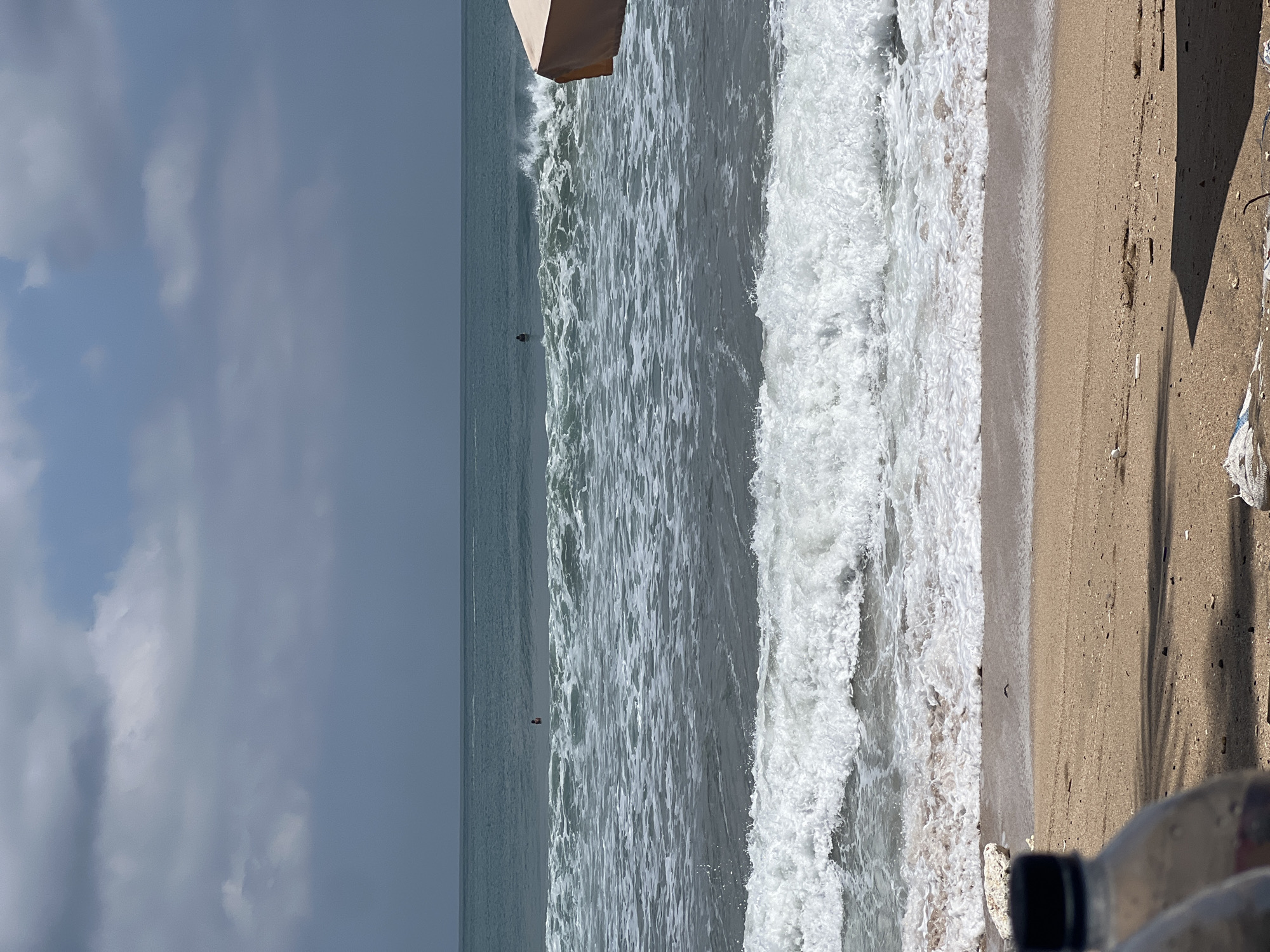 A wide shot captures a beach scene in Kuta, Indonesia. The ocean stretches to the horizon, with a grey sky overhead. Waves are breaking on the shore, with white foam and spray visible. Two people are in the water, appearing to be swimming or surfing. The sandy beach is in the foreground, with some debris and two plastic bottles visible. A portion of a beach umbrella is visible on the right side of the image. The overall scene depicts a cloudy day at the beach.