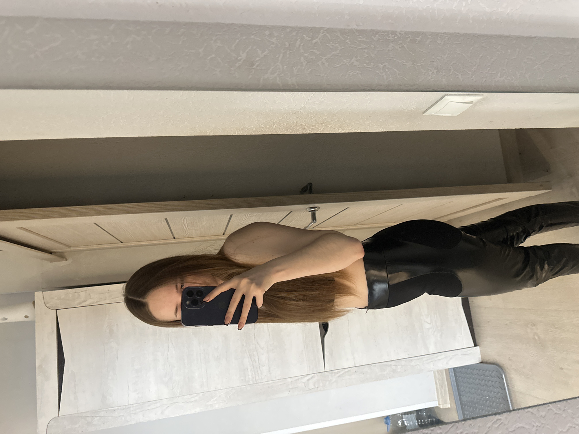 A woman stands in an indoor setting, taking a selfie in a mirror. She is wearing black leggings. The background features a countertop and shelves.