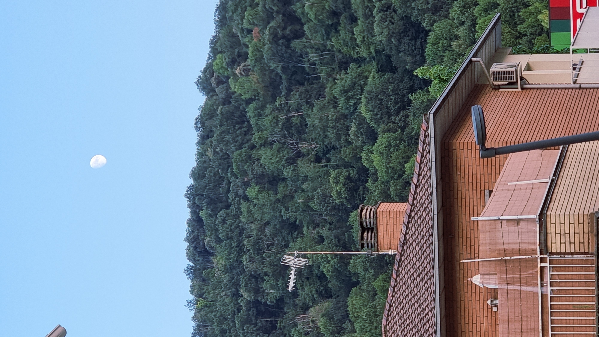 The image depicts a view from a residential building towards a densely forested hillside under a clear blue sky. The moon, appearing as a waning crescent, is visible in the upper portion of the sky. In the foreground, the terracotta tiled roof of a brick house is prominent. A chimney made of reddish-brown bricks, topped with two circular vents, stands on the roof. An antenna array is mounted on a pole next to the chimney. The side of the house features a balcony with white railings and a beige-colored privacy screen. An air conditioning unit is mounted on the exterior wall of the building. The background is dominated by a lush green hillside covered in a thick canopy of trees, suggesting a natural or semi-rural setting within the municipality of Sant Joan Les Fonts, Spain. No people are visible in the frame.
