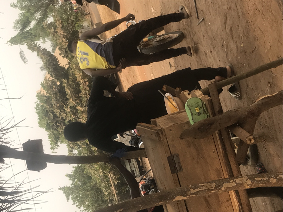 Two men are outside on a sandy surface. One man, wearing a yellow jersey with "31" on the back and black pants, stands near a bicycle wheel. The other, dressed in black, appears to be working near a wooden structure, possibly a cart. A green object and a yellow container sit on top of the wooden structure. The scene includes trees and foliage in the background.