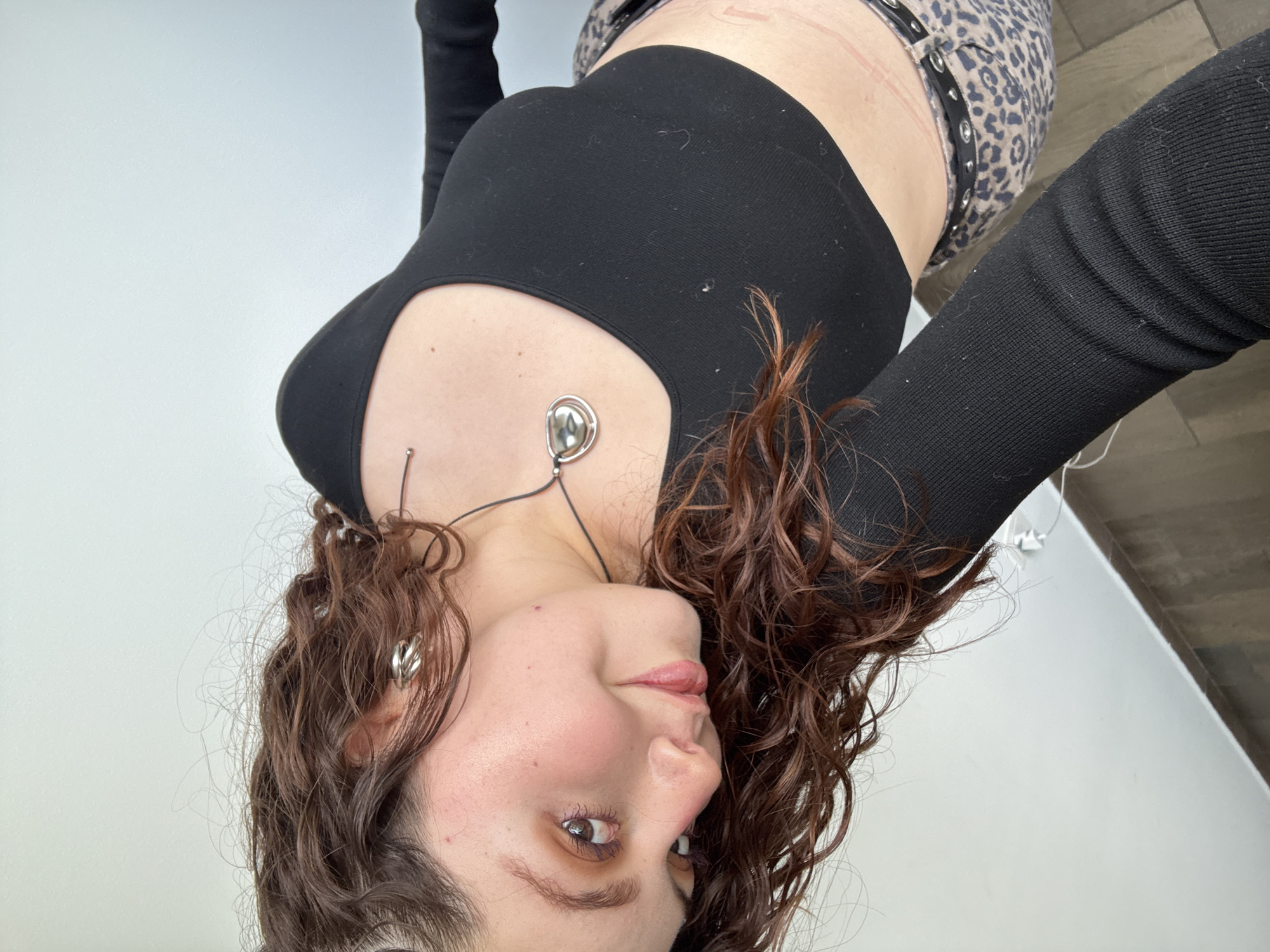 A young woman poses for a selfie. She is facing the camera from an upside-down angle. She has brown curly hair, fair skin, and is wearing a black long-sleeved crop top. She has multiple piercings, including one near her collarbone and in her ear. A silver necklace is around her neck. She is wearing leopard-print pants. The background appears to be a plain white wall and a glimpse of a light-colored floor. The image does not provide any context about being in Mexico City.