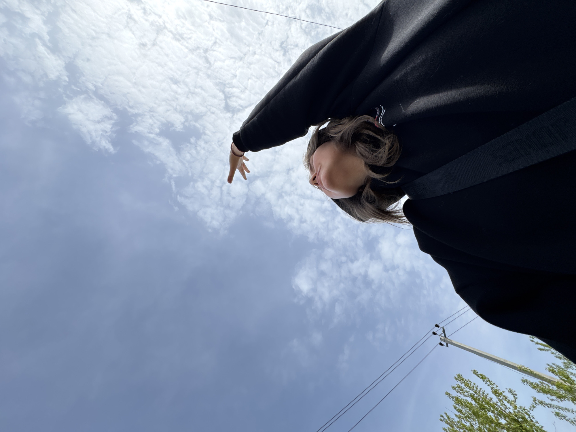 A low-angle photograph shows a person with short, wavy brown hair, wearing a black hooded sweatshirt and a black strap across their chest, extending their left hand upwards against a partly cloudy sky. The scene includes power lines and a light green tree in the lower right corner. The location is Горный, Russia.