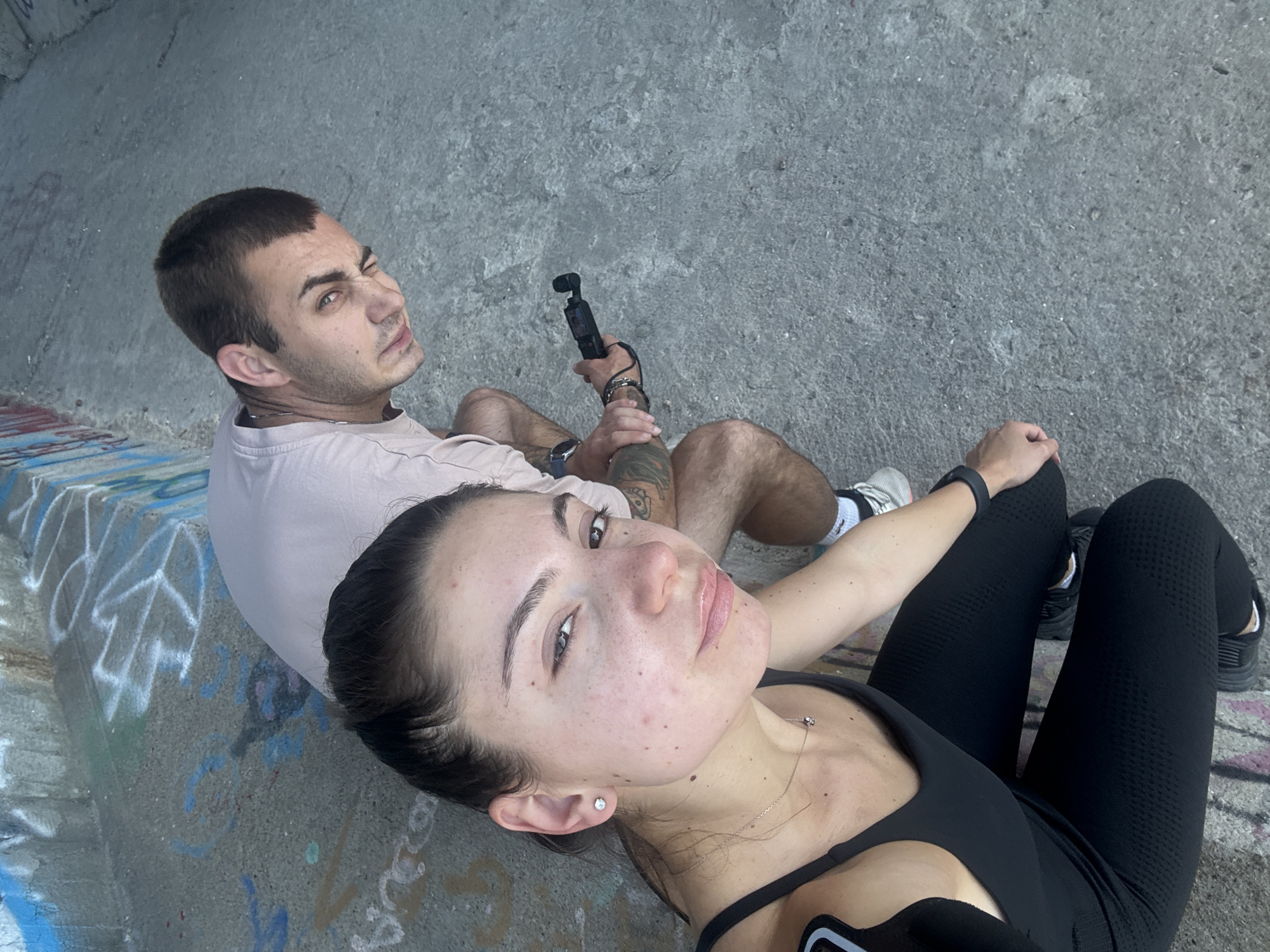 Two individuals are crouched on a concrete surface marked with graffiti. The person in the foreground, a woman with dark hair pulled back, is looking towards the camera and wearing a black tank top and leggings. She has a necklace with a small pendant. The second person, a man with short brown hair, is positioned behind and to her right. He is wearing a light-colored t-shirt and shorts and is holding a small black electronic device. The background shows a concrete wall with blue and white graffiti. The overall environment suggests an urban outdoor setting, potentially an underpass or abandoned structure, common in areas like Varna, Bulgaria.