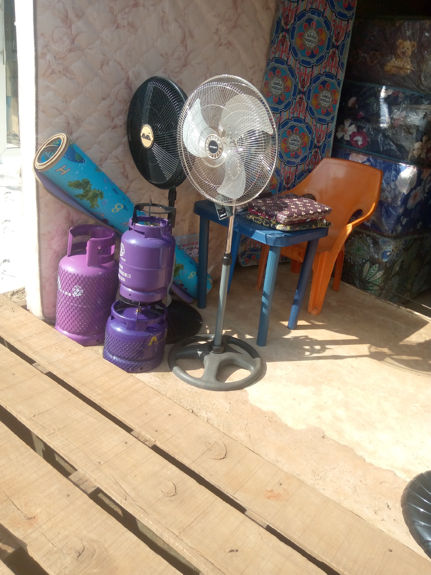 In Nsugpe, Nigeria, a scene unfolds showcasing an outdoor storage area near a building. Dominating the foreground are several purple gas cylinders, one large and two smaller ones stacked on top of each other, all fitted with black netting. To the left, a rolled-up blue mat with cartoon imagery is leaning against a wall. Behind the gas cylinders and mat, a large, patterned mattress with a quilted texture is propped up. Further back, two electric fans are visible; one is a black, standing fan with a circular base, and the other is a silver, oscillating fan with a circular grille, also on a stand. A small blue table supports a stack of colorful folded cloths or small cushions. An orange plastic chair is positioned behind the table. In the background, more stacked mattresses or similar items, some wrapped in clear plastic, line the wall of the structure, which appears to be a shop or storage space. The ground is a mix of concrete and dirt, with wooden planks laid out in the immediate foreground. Sunlight casts distinct shadows across the scene, indicating it is daytime.