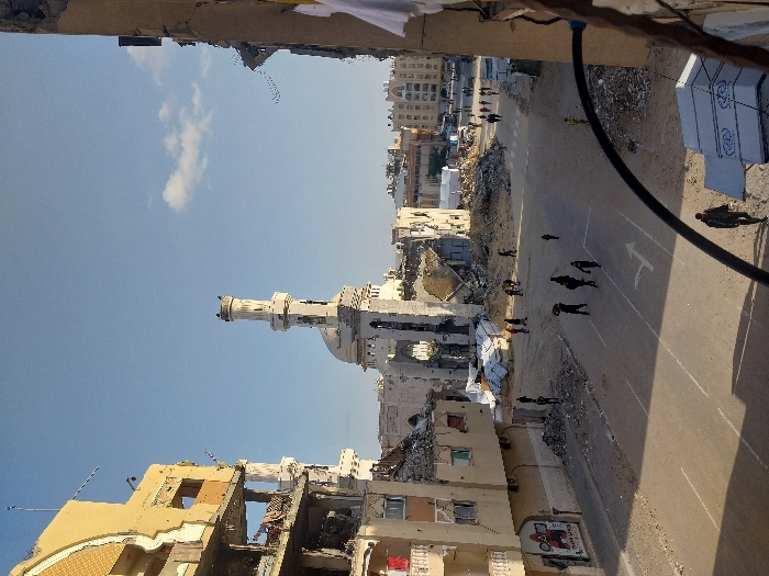 The image depicts a severely damaged urban environment in Khan Yunis, Palestinian Territory, under a clear blue sky with a few sparse clouds. In the center, a prominent mosque with a tall, slender minaret and a dome shows extensive structural damage; large sections of its walls are destroyed, revealing the interior, and significant rubble surrounds its base. To the left, a multi-story, light-colored building is heavily damaged, with multiple floors exposed, missing exterior walls, and visible rebar; graffiti featuring red text and figures is visible on its lower facade. A partial view of another extensively damaged building frames the right side, with white laundry hanging from its visible structure.
A paved road, marked with white lane lines and a directional arrow, runs through the foreground and mid-ground. The areas surrounding the mosque and adjacent to the road are covered in large piles of construction debris, including concrete chunks and twisted materials. Approximately 15-20 individuals, dressed in civilian clothing, are scattered across the scene, walking on the road and navigating the rubble-strewn areas. Some individuals appear to be carrying objects. A black curved pipe or cable crosses the lower right foreground, partially obscuring a white corrugated structure below it. The scene is illuminated by bright sunlight, casting distinct shadows from people and structures.