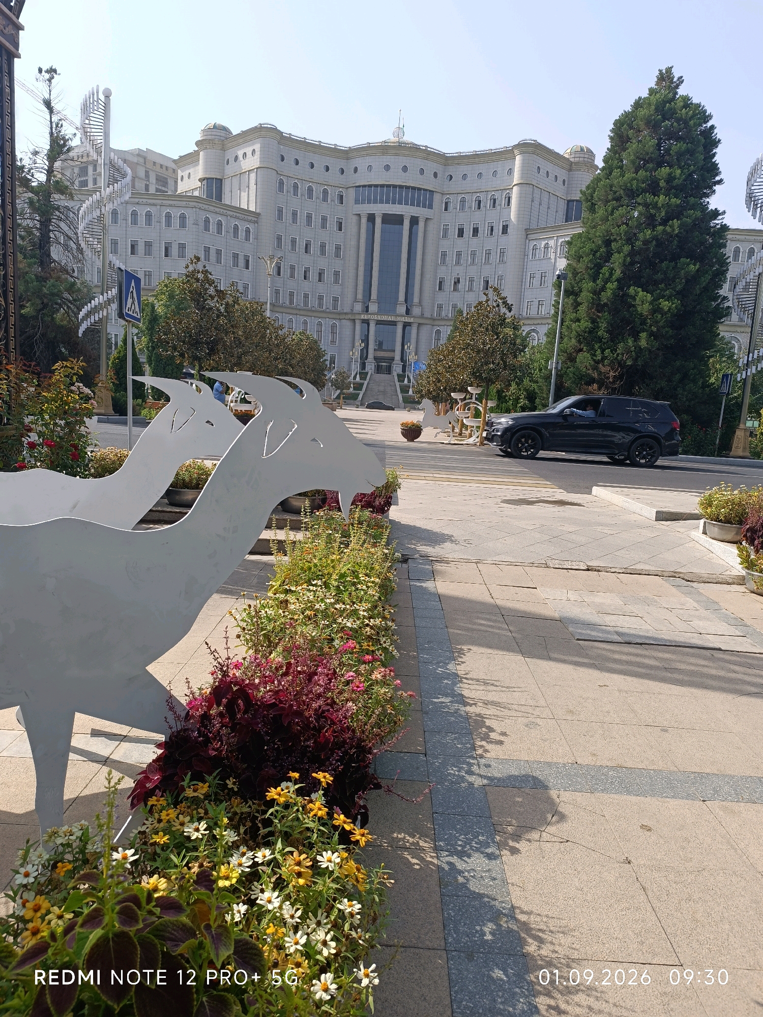 The image shows a wide, paved street in Dushanbe, Tajikistan, in front of a large, modern, light-colored building with many windows and decorative columns. A black SUV is driving on the street, and a pedestrian crossing sign is visible. In the foreground, there are flower beds with colorful flowers and decorative sculptures of white, stylized animals, possibly horses or goats. The sky is clear and bright, suggesting a sunny day. A watermark in the lower left corner reads "REDMI NOTE 12 PRO+ 5G", and in the lower right corner, a date and time stamp reads "01.09.2026 09:30". The architecture of the building and the presence of the sculptures suggest this is a public space, possibly a government building or a cultural institution.