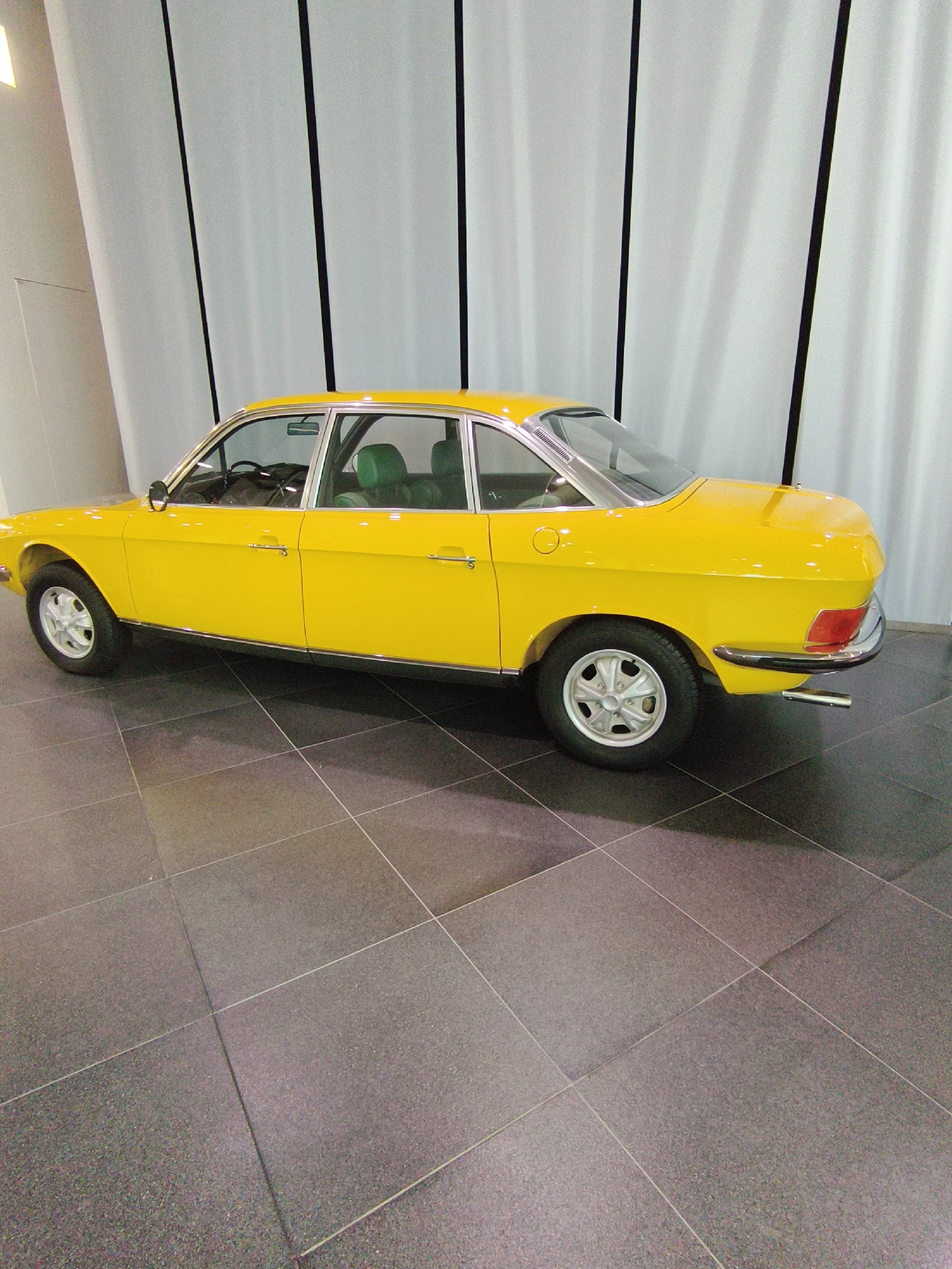 A bright yellow, vintage sedan is parked on a tiled floor indoors. The car is viewed from a side angle, showing its left profile. It has a classic design with chrome accents on the door handles, window trim, and bumpers. The interior appears to have green upholstery visible through the windows. The background consists of large, white, vertically pleated curtains and a light-colored wall with a curved architectural feature. The floor is made of dark grey square tiles with visible grout lines. There are no people or animals in the image. This scene is likely within the Audi Museum Mobile in Ingolstadt, Germany, given the presence of vintage automobiles and the modern architectural style.