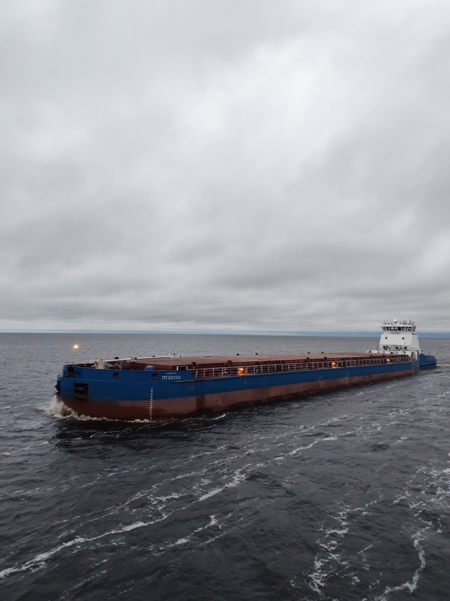 A barge, with blue and brown hull and white superstructure, traverses dark water under a gray, overcast sky. The vessel, marked "IT 82684," leaves a wake. The environment suggests a body of water near a shoreline; the scene possibly depicts a location near Bereg, Russia.