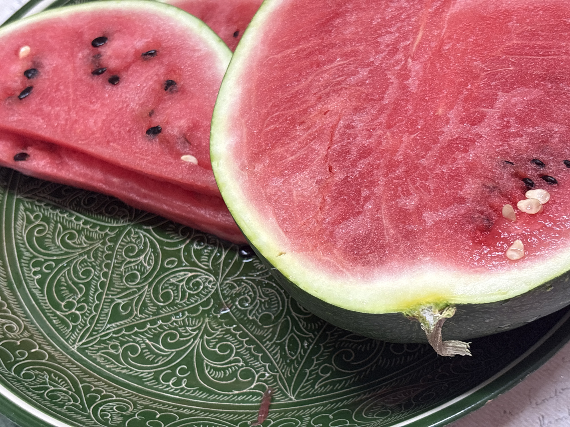 A close-up shot shows a cut watermelon on an ornate green plate. The watermelon is bright pink and has black seeds. Two slices of watermelon are behind the half-melon. The plate has intricate white designs. The location is presumed to be Lukintsy, Russia.
