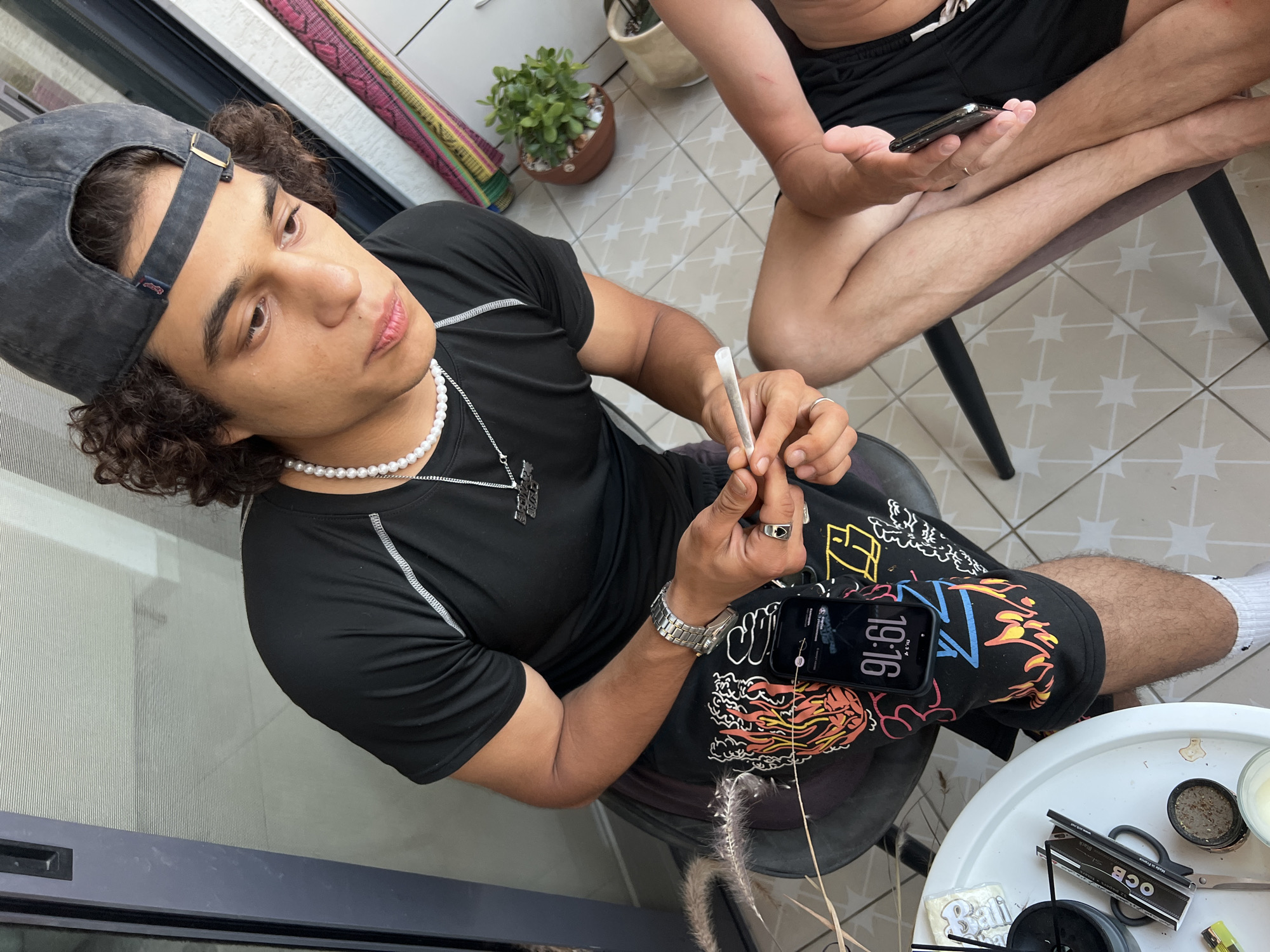 The image depicts two individuals, likely young men, engaged in activity outdoors. The foreground features a young man wearing a black t-shirt, a pearl necklace, a silver watch, and dark patterned pants. He has curly dark hair and is wearing a dark baseball cap. He is holding a partially rolled cigarette or joint between his fingers, preparing to smoke. A smartphone is attached to his pants pocket, displaying the time "19:16". To his left, a white table holds various items, including what appears to be a grinder with cannabis residue, scissors, a pack of rolling papers labeled "OCB", and a decorative item. The background shows a tiled floor, a potted plant, and a balcony railing with some colorful fabric draped over it. The presence of the OCB rolling papers and the grinder with cannabis residue suggests the activity involves the preparation of a cannabis cigarette. The overall setting appears to be a residential balcony or outdoor seating area. There is no explicit visual information that places this scene specifically in Kiryat Bialik, Israel.