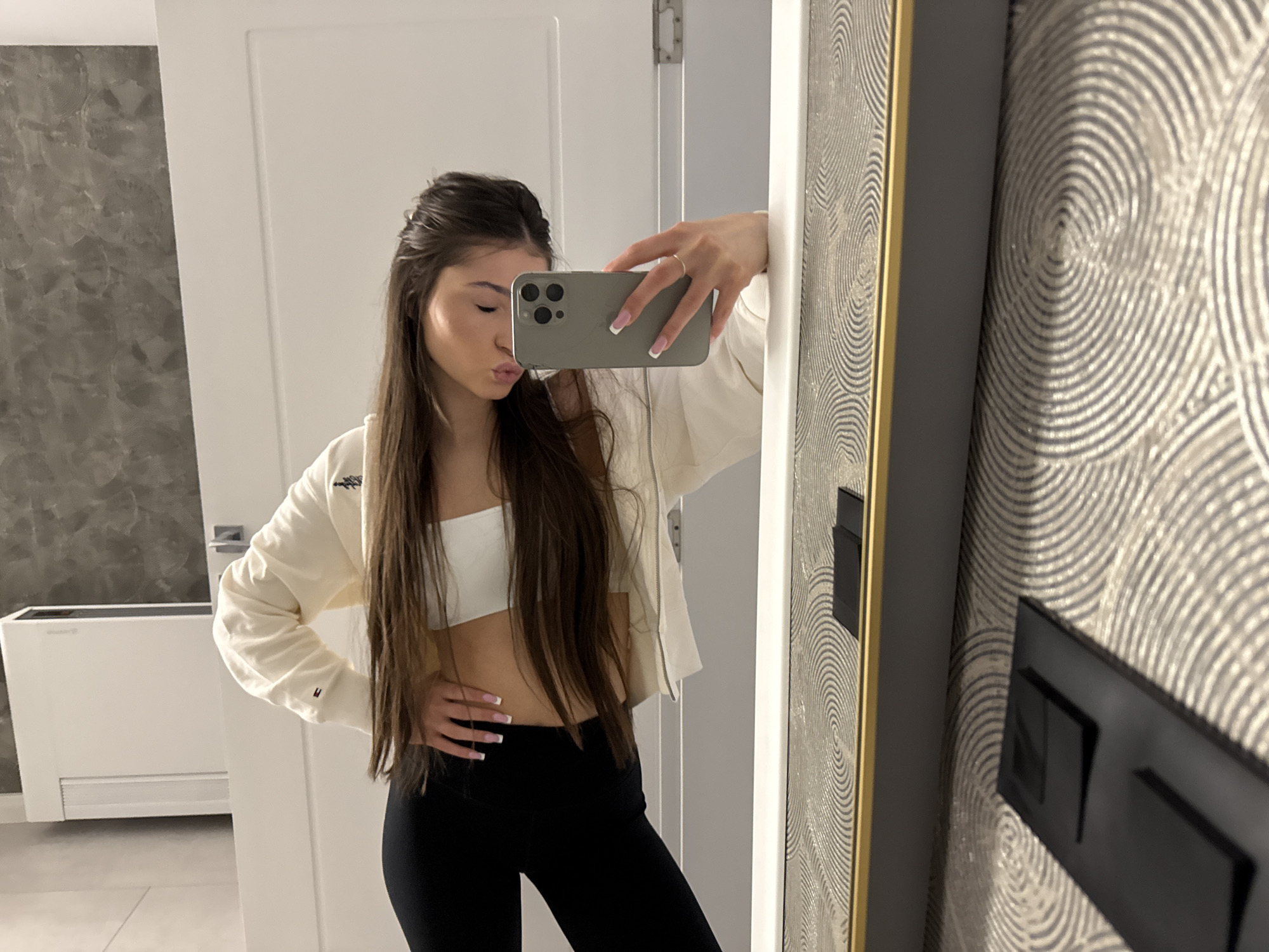 A young woman poses for a selfie in front of a mirror. She is wearing a white tube top, black leggings, and an off-white jacket. The background includes a white door, a gray-textured wall, a white heating appliance, and a patterned wallpaper in a mirror frame. The location is likely indoors, possibly in a home or apartment. The city is Plovdiv, Bulgaria.