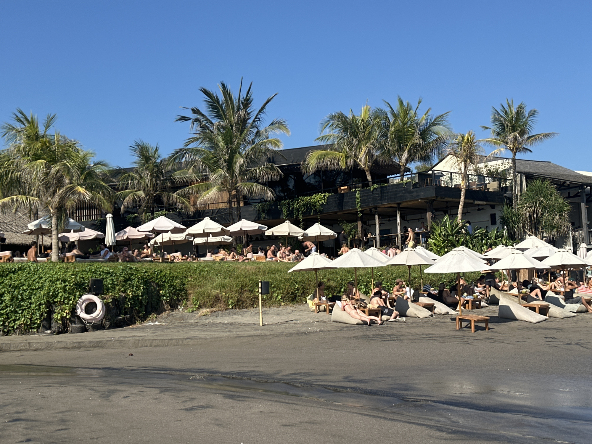 The image presents a detailed view of a beach club scene in North Kuta, Indonesia, under a clear blue sky. In the background, a multi-story building with dark roofs, open architectural elements, and numerous balconies is partially obscured by lush green palm trees and dense foliage. A prominent green, vegetated bank separates the building's lower level from the beach. On the left side of this bank, a white life preserver with a red band is mounted on a dark post. The midground and foreground consist of a dark, sandy beach. Approximately 20 individuals, primarily adults, are distributed across the sandy area, engaged in leisure activities. They are seated on light-colored beanbag chairs, wooden lounge chairs, and low wooden tables, all positioned beneath numerous large, light-colored beach umbrellas. A dark sandy path or road occupies the immediate foreground.