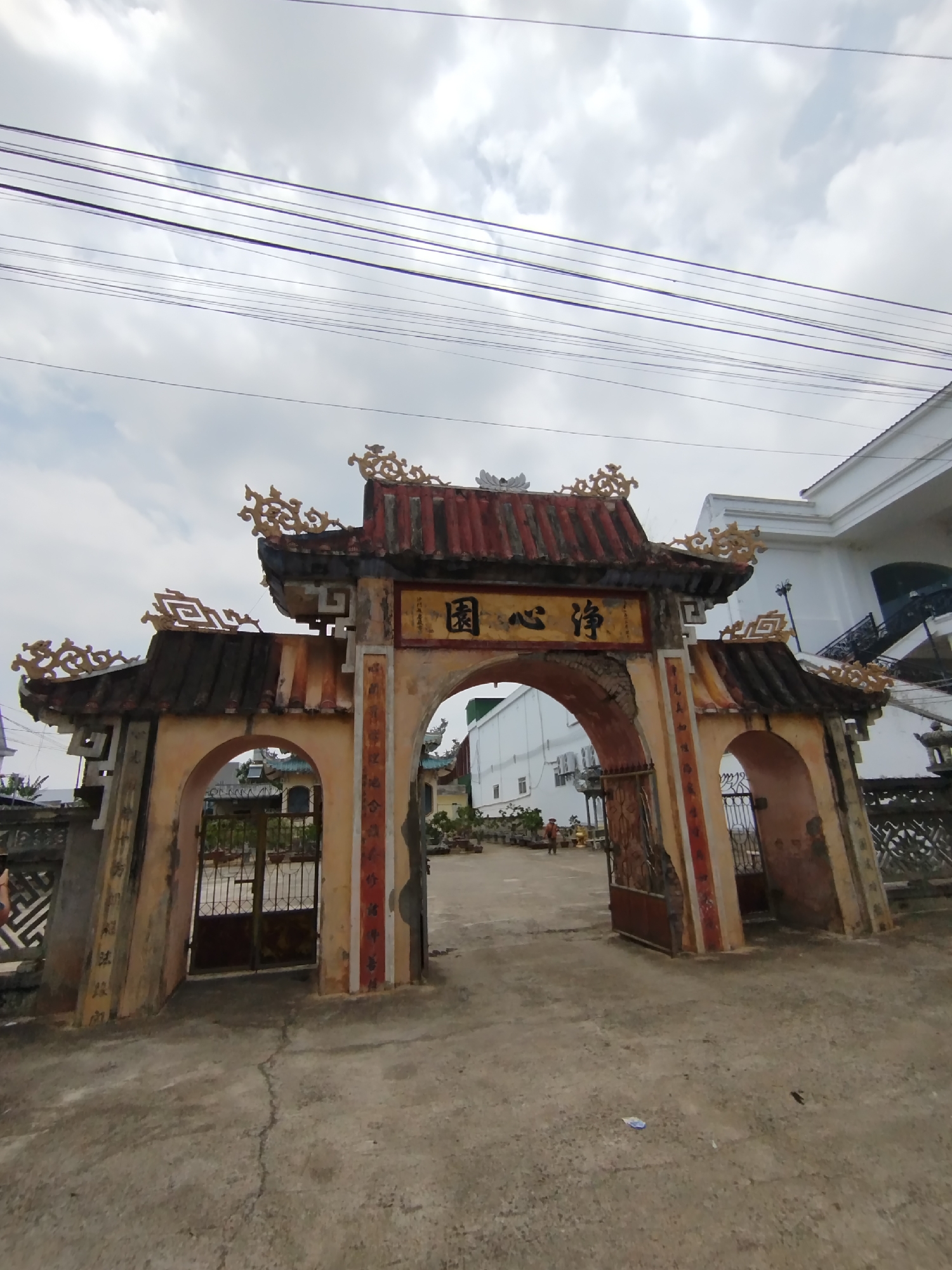 The image shows an ornate, weathered gate, likely the entrance to a temple or religious site. The gate features a central archway with smaller arched openings on either side, all constructed of aged stucco or plaster and decorated with traditional East Asian architectural elements. A red-tiled roof tops the structure. A sign with Chinese characters is prominently displayed above the main arch. The pavement leading up to the gate is cracked and aged, suggesting a well-traveled area. In the background are buildings, including a white two-story structure and what appears to be a smaller, more traditional building. Power lines stretch across the top of the frame, indicating an urban or suburban location. The image was likely taken in Phú Lộc, Vietnam.