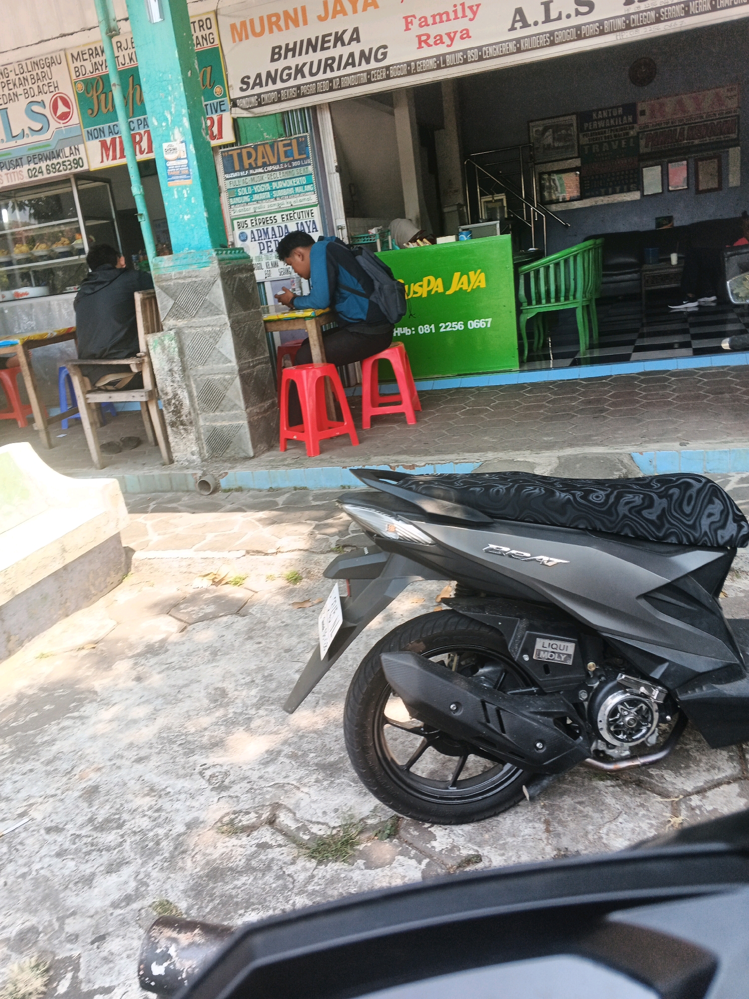 The image depicts a street scene in Ungaran, Indonesia, likely in front of a business establishment. In the foreground, a dark-colored motorcycle is parked on a concrete ground with irregularly shaped paving stones. The motorcycle has a patterned seat cover and a "LIQUI MOLY" sticker on its engine.
Behind the motorcycle, a man wearing a dark jacket and backpack is seated at a yellow table, looking down at something he holds in his hands. He is sitting on a red stool. Another person, wearing a dark jacket, is seated at a similar table with a blue stool in the background, facing away from the camera.
The background features several signs. A large green sign with "GUSPA JAYA" in yellow lettering and a phone number is prominently displayed. Above this, other signs are visible, including "TRAVEL" and company names like "MUNRI JAYA," "BHINEKA," "SANGKURIANG," and "A.L.S." A green chair is also visible near the business entrance. The building appears to be a commercial establishment, possibly a travel agency or bus station. The overall environment suggests a busy roadside location.