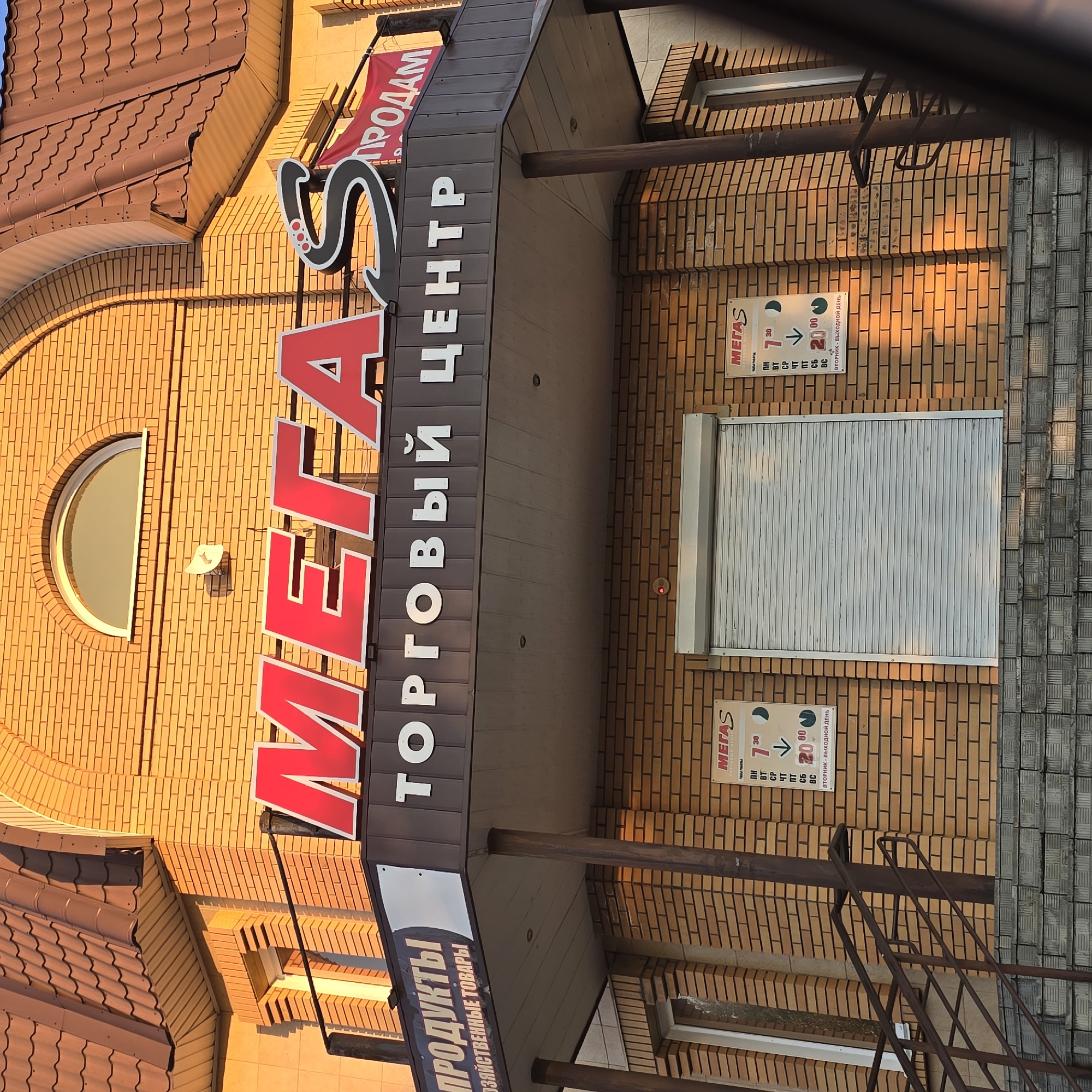 The image shows the storefront of a shopping center named "MEGAS" located in the city of Belokurikha, Russia. The building is constructed with orange brick. Above the entrance, large red letters spell out "MEGAS" followed by a stylized "S". Below this, in white letters, is the text "ТОРГОВЫЙ ЦЕНТР" (Shopping Center). A red banner to the right of the main sign reads "ПРОДАМ" (For Sale). On the brick wall to the left and right of the entrance, there are signs displaying the operating hours for "MEGAS". The entrance itself is covered by a closed, light-colored roller shutter. The scene is illuminated by warm, golden sunlight, likely from a sunrise or sunset, casting long shadows. Stairs lead up to the entrance.