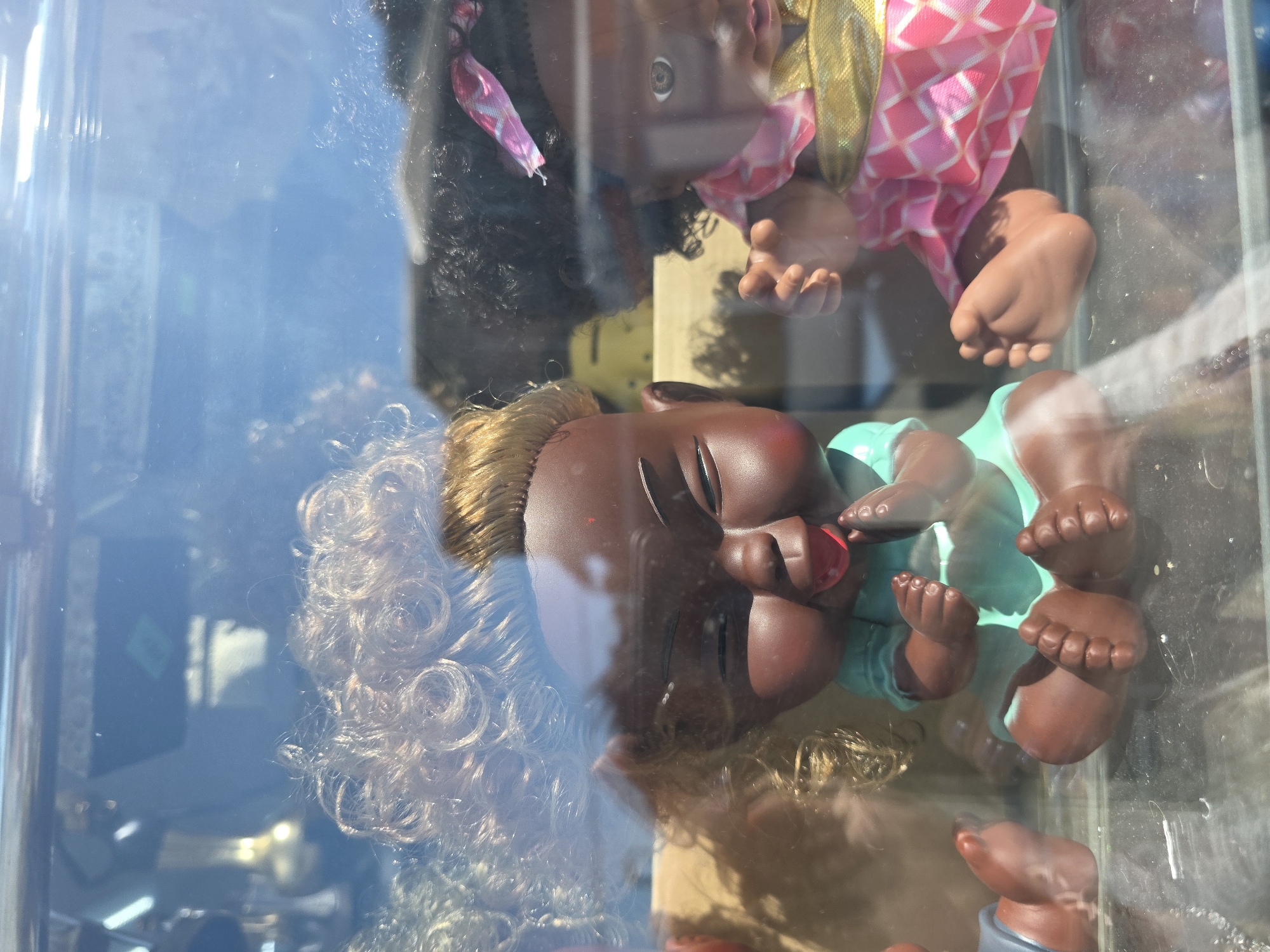 This image, captured in Sochi, Russia, presents two dark-skinned baby dolls positioned behind a reflective glass surface, likely within a display case. The leftmost doll features a dark brown complexion with pronounced cheeks, closed eyes represented by upward slants, and bright red lips. It wears a light blue-green top and has short, light blonde hair. Its hands are clasped together in front of its chest, and its bare feet rest on a clear surface. A significant reflection of voluminous curly blonde hair on the glass partially overlays this doll and the upper background. To its right, a second dark-skinned doll is visible, wearing a pink patterned dress with a gold sash and dark, curly hair adorned with a pink and white patterned ribbon. One brown eye is visible, and its right hand is slightly raised. Both dolls are in a seated posture. The background, seen through and reflected on the glass, appears as a blurred blend of blue sky and indistinct reflections of internal objects, including a golden, decorative item in the upper left. The glass surface exhibits minor smudges and reflections of light sources.