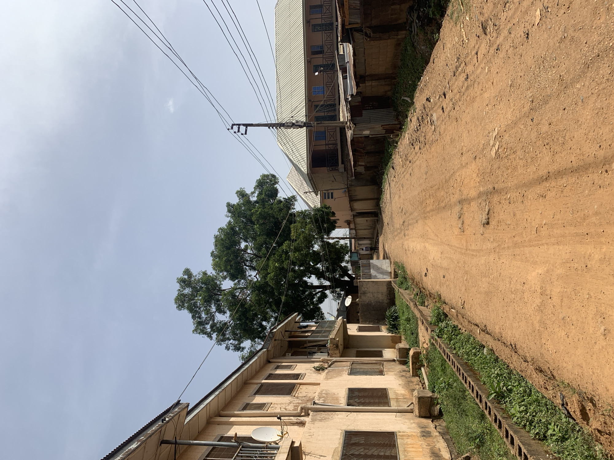 A daytime shot reveals a narrow, unpaved street in Rafin Par, Nigeria. Buildings flank either side of the street, one taller and more modern with a light-colored metal roof, and the other appearing older with a weathered facade and multiple windows. A large tree stands between the buildings. Power lines stretch across the sky above the buildings. A concrete drainage ditch runs alongside the street. There are no people visible in the image.