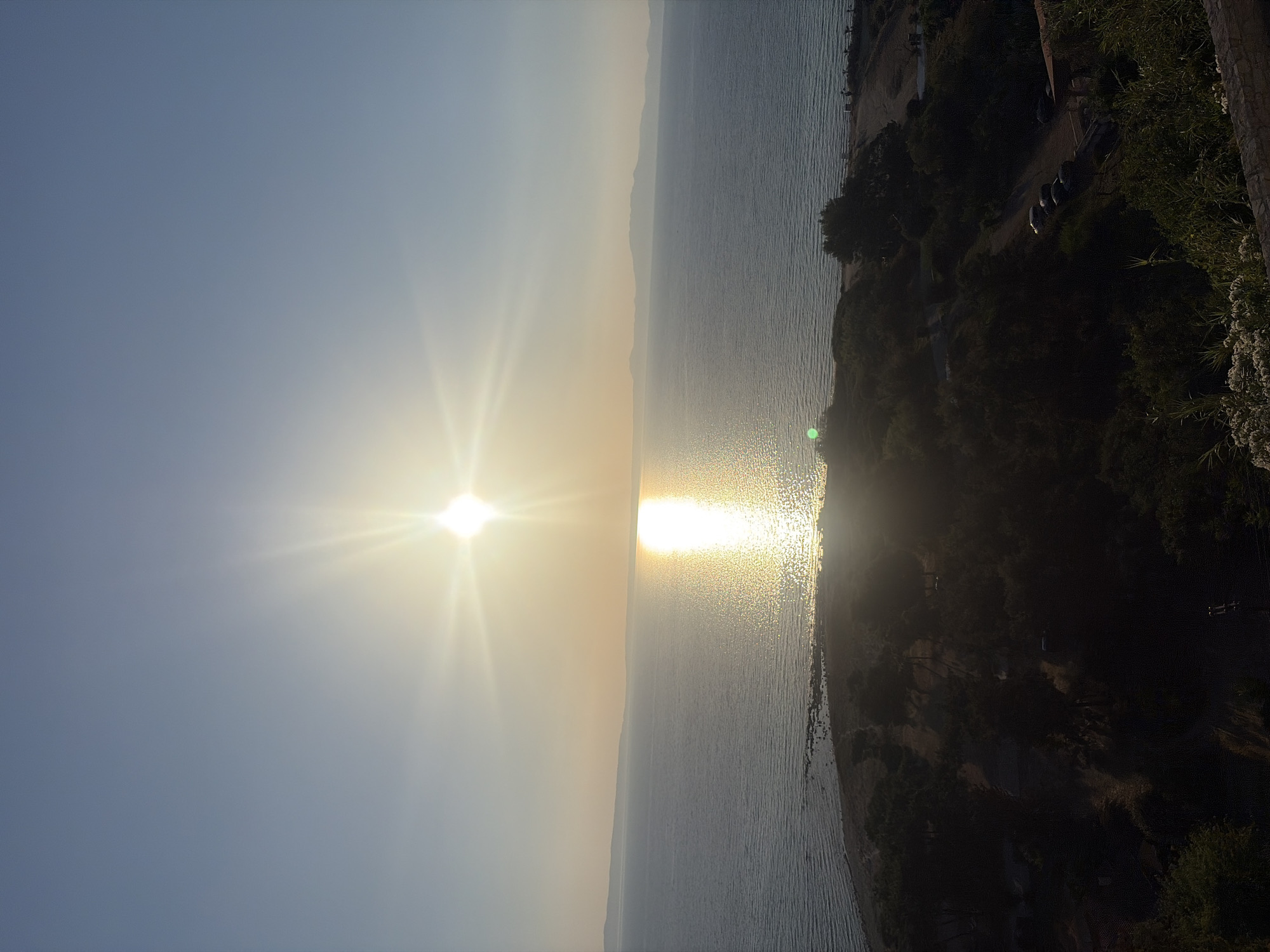 This is a landscape photograph taken from an elevated viewpoint overlooking the sea and a coastal area. The sun is high in the sky, slightly to the left of the center, casting a bright glare and a shimmering reflection on the water. The sea stretches out to the horizon, meeting a hazy, mountainous coastline in the distance. In the foreground, there is a densely vegetated hillside with trees and shrubs. A dark dirt path winds through the vegetation, with several cars parked along its side. The overall impression is one of a tranquil coastal scene, possibly during sunset or sunrise, with the sun's intense light dominating the view. The location is identified as the Kassandra Municipal Unit in Greece.