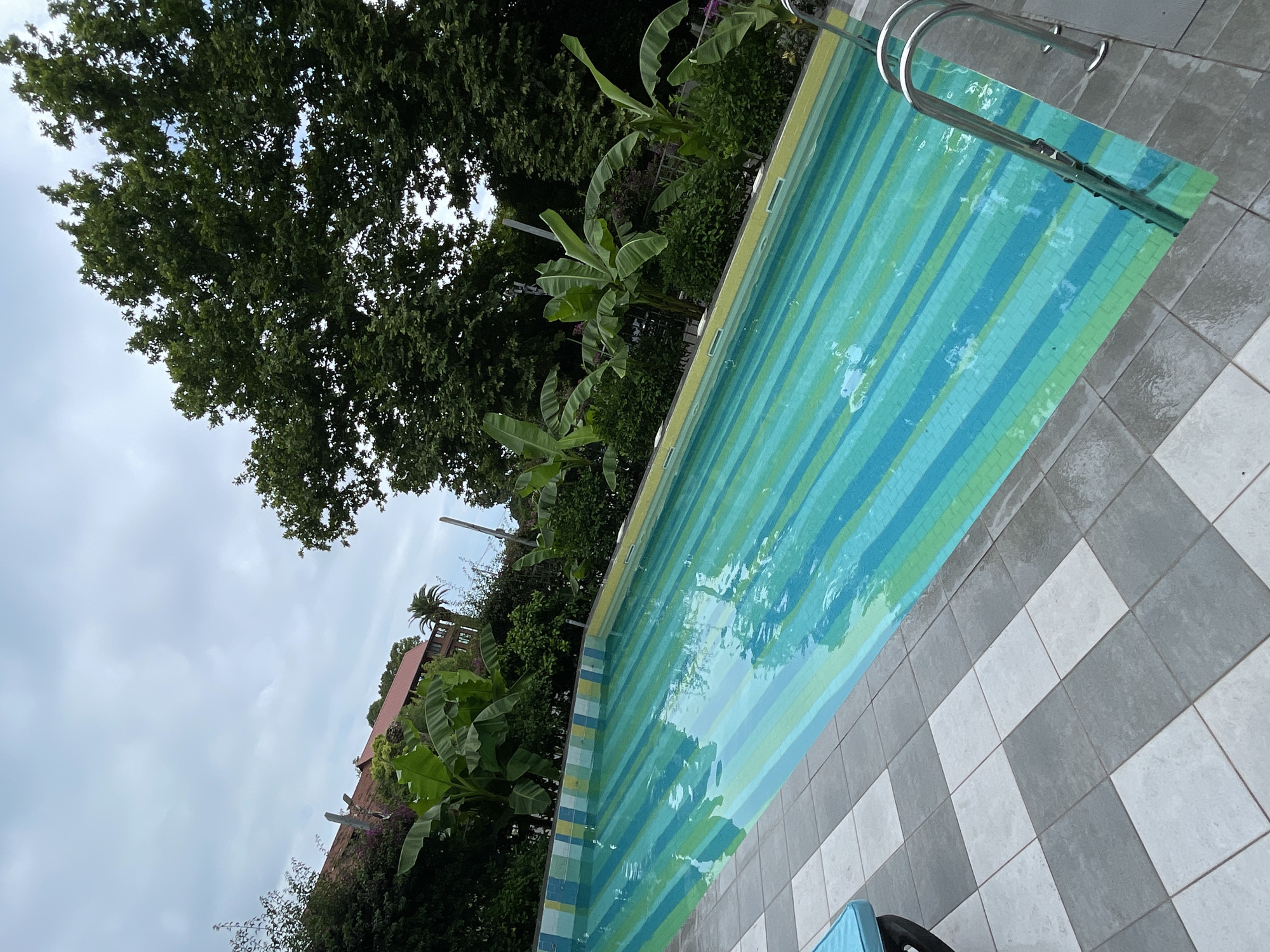 The image shows an outdoor swimming pool with a striped pattern in shades of green and blue. The pool is surrounded by a tiled patio with a checkerboard pattern of gray and white squares. Several banana trees and other lush green vegetation are visible behind the pool. In the background, a building with a red tiled roof can be seen. The sky is overcast. The scene appears to be a resort or a private residence in Abaata, Abkhazia. There are no people visible in the image, nor any actions or interactions taking place.