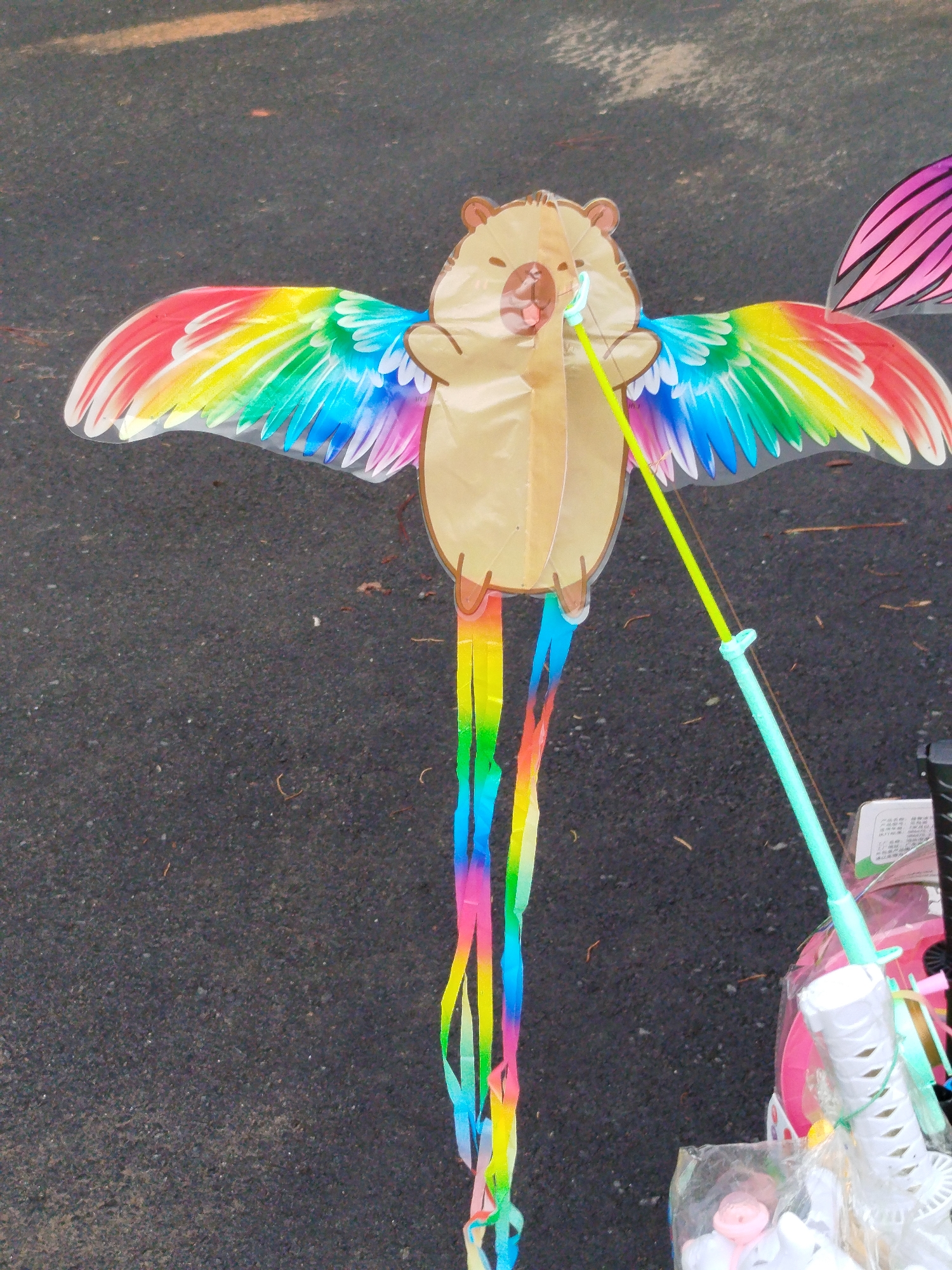 A kite with a cartoon squirrel head and rainbow-colored wings and tail streamers is laying on asphalt. The kite is attached to a green plastic handle and reel. Other unidentified objects in plastic packaging and pink toys are visible to the right of the kite. The image was taken during daylight hours. The location is unknown.