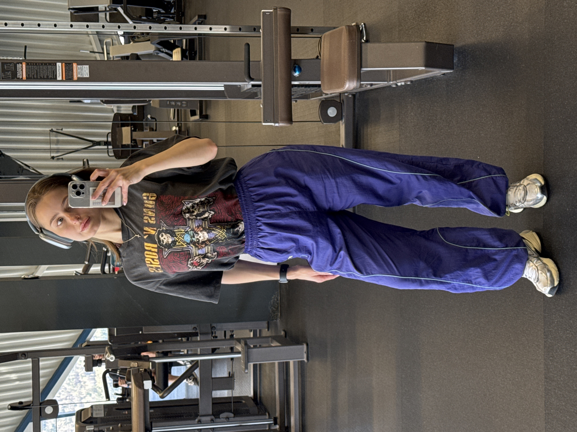 A young woman is taking a selfie in a gym. She is wearing headphones, a black t-shirt with a Guns N' Roses graphic, blue sweatpants with a light blue stripe, and white sneakers. She is holding a smartphone with a gray case. The background includes gym equipment, such as a rowing machine and cable machines. The image does not provide any information about the city of Krasnoyarsk, Russia.
