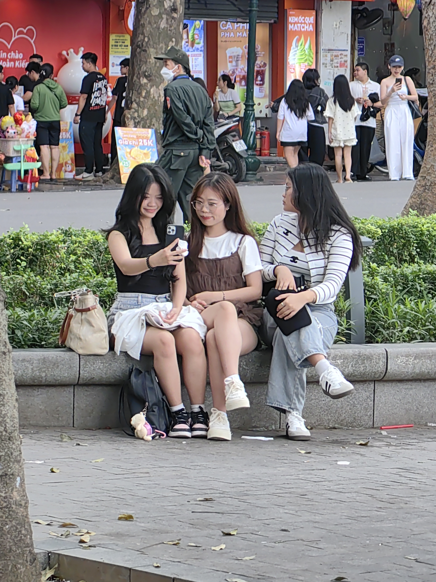 Three young women are sitting on a stone bench alongside a sidewalk in Hà Nội, Vietnam. The woman on the left is taking a selfie with her phone, and the other two are looking at the phone. All three are dressed casually. Behind them is a green hedge. Behind the hedge are several pedestrians and a police officer, and behind the pedestrians are several stores. Signage for "Kem Oc Que Matcha" and "Cafe Pha May" is visible. A sign advertising "Can Yang 9990" for 25,000 dong is visible in the lower left.