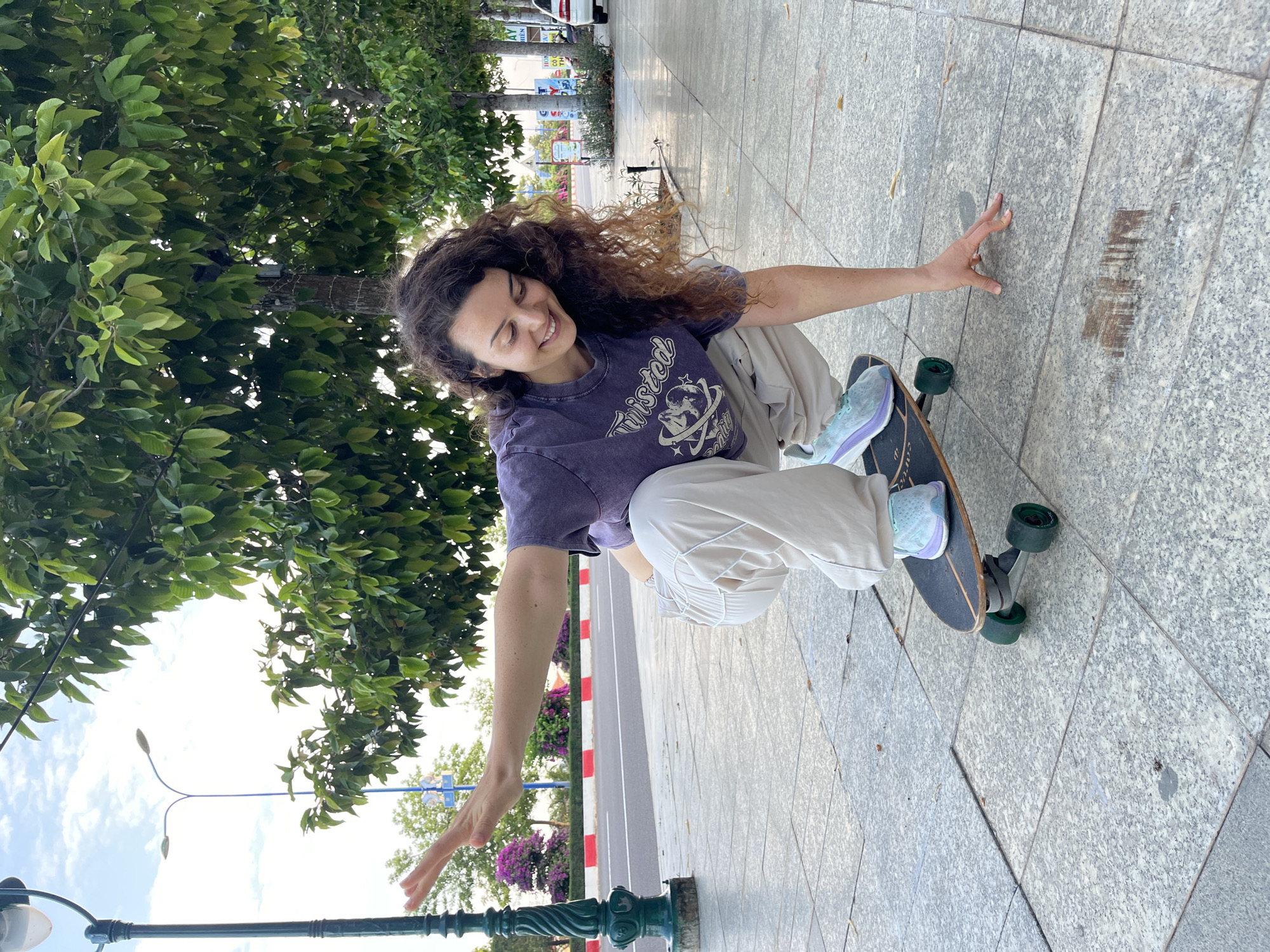 A young woman with curly brown hair is crouching on a skateboard on a paved sidewalk. She is wearing a purple t-shirt with a graphic and light-colored baggy pants. Her right hand is touching the ground, and her left arm is extended outwards. The skateboard has green wheels and a black deck. In the background, there are trees, streetlights, a road with a red and white striped median, and some buildings. The scene appears to be outdoors in a city, possibly Phu Thuy Ward, Vietnam, indicated by signage in Vietnamese visible in the background. The lighting suggests it is daytime.