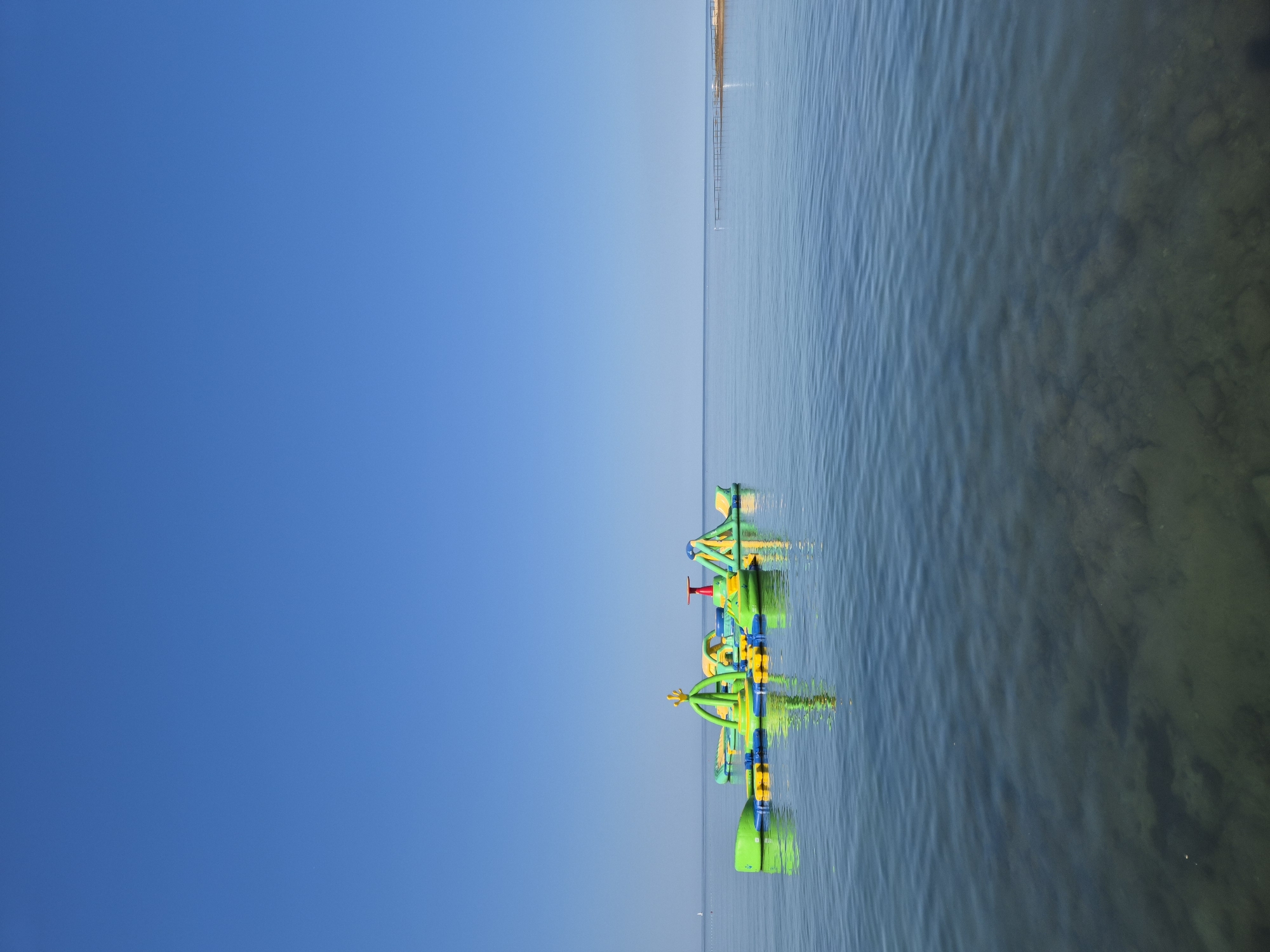 The image depicts a tranquil scene of an inflatable water park floating on the calm, clear waters of the Adriatic Sea near Katoro, Croatia. The sky is a vibrant, cloudless blue, meeting the horizon in a sharp line. The water is transparent enough to reveal the rocky seabed beneath, with gentle ripples distorting the reflections of the inflatable structures. The water park itself is a collection of brightly colored, interconnected inflatables in shades of green, yellow, and blue, featuring various slides, climbing structures, and platforms. In the distance, a long wooden pier extends into the sea, and a small sailboat is visible on the far left horizon. The overall impression is one of a serene summer day, with the water park set against the expansive, peaceful backdrop of the sea and sky.