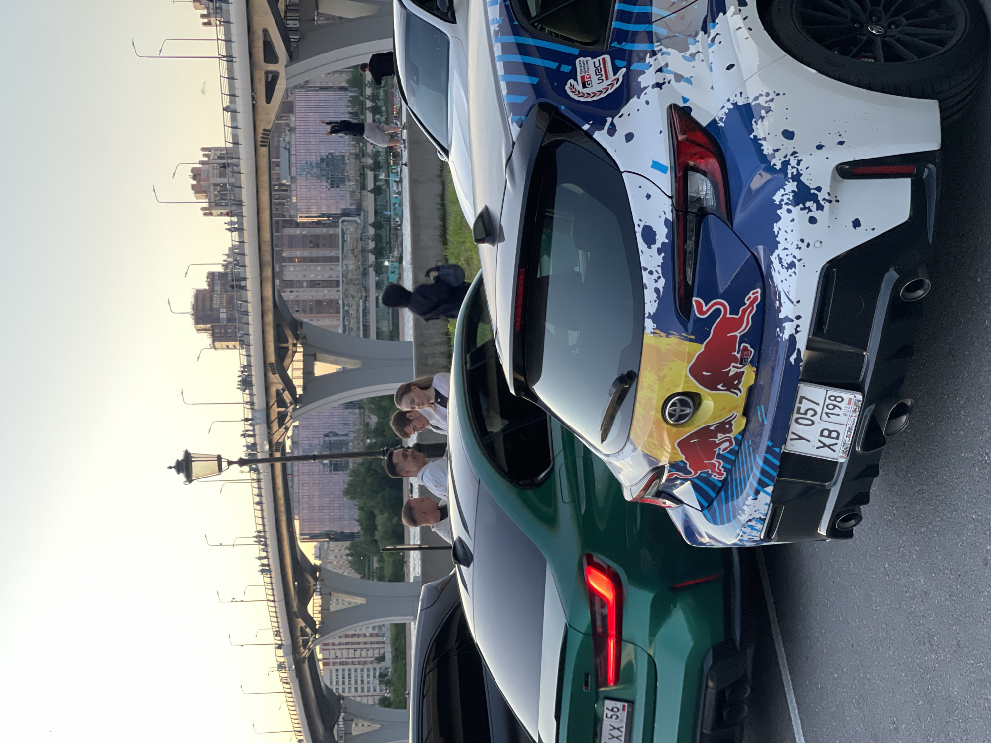 The image shows a group of people standing near cars with a bridge in the background. In the foreground, there are two cars: a green BMW and a white car with a colorful Red Bull livery. The green BMW has its rear end facing the camera, with its taillights illuminated. The white car's rear is also visible, showcasing a dynamic paint scheme. Four people are standing between the two cars, engaged in conversation. One man is smiling and looking at the others. In the middle ground, a bridge spans across a body of water, with pedestrians walking along it. The sky is clear and appears to be sunset. The environment suggests an urban setting, possibly a waterfront area.