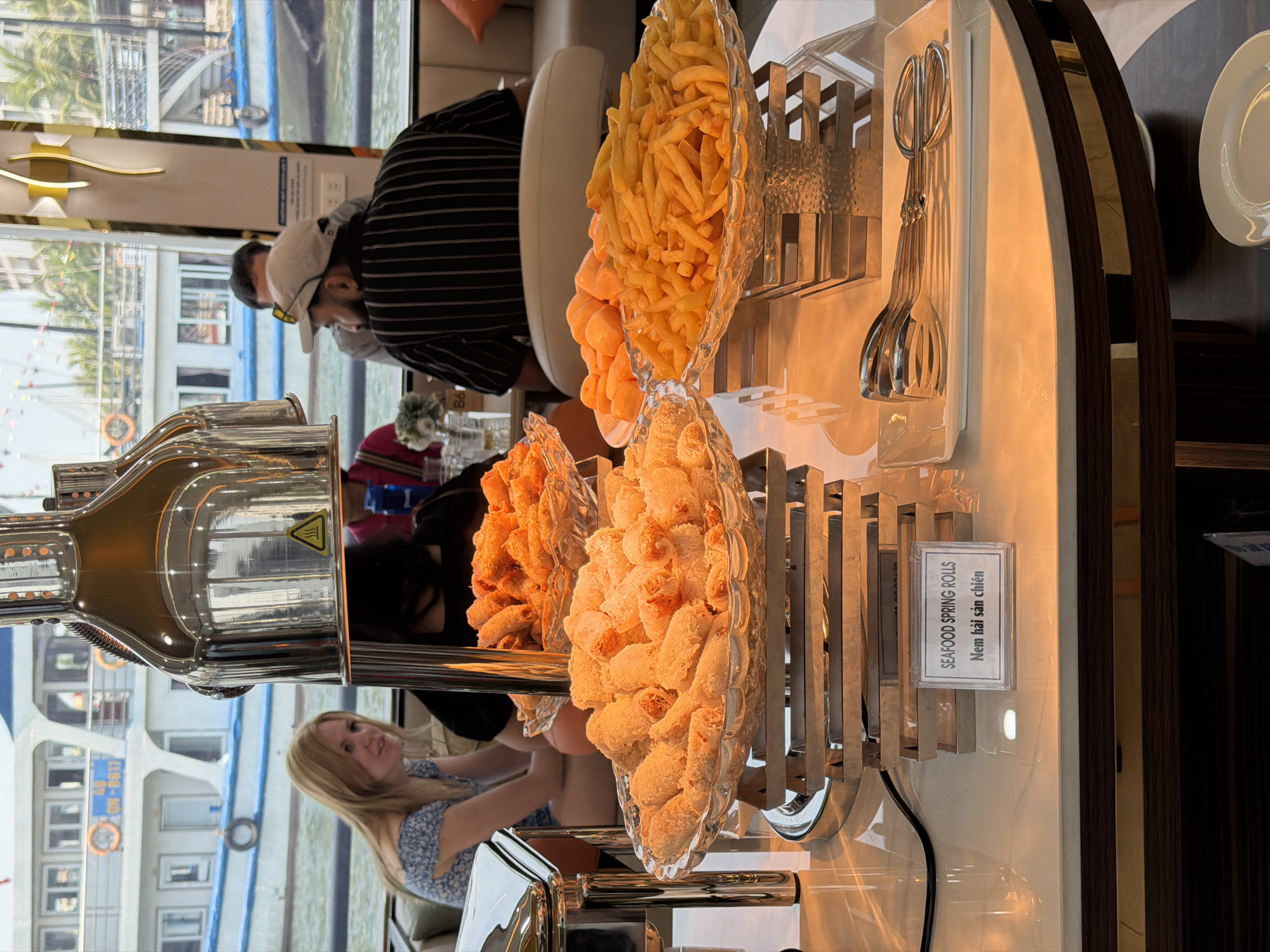This image depicts a buffet setup inside what appears to be a boat or cruise ship, offering a view of the outside environment. The photograph is oriented vertically, rotated 90 degrees clockwise.
In the foreground and mid-ground, a selection of fried foods is displayed in multiple clear, decorative glass dishes. These include a large quantity of golden-brown french fries, a bowl of bright orange, puffed snacks, and two bowls filled with golden-brown, cylindrical items. Adjacent to these, a small white sign clearly labels them as "SEAFOOD SPRING ROLLS" and "Nem hải sản chiên" (Vietnamese for "Fried seafood spring rolls"). Metal serving tongs are neatly arranged on a rack next to the food, ready for use. The food is kept warm in polished metal chafing dishes, one of which features a yellow warning symbol for heat.
In the background, several individuals are present. A man wearing a striped shirt, a white baseball cap, and sunglasses pushed up is seated. To the lower left, a woman with blonde hair, wearing a patterned dress, is also seated, looking towards the right. The setting appears to be a comfortable dining area with large windows.
Beyond the interior, a bright, sunny daytime scene unfolds. The windows offer a clear view of a body of water, with other boats visible, including a prominent white vessel with blue trim and Vietnamese text and numbers (partially legible) on its side. Palm trees and distant land can also be seen, indicating a scenic coastal or island location. The scene suggests a relaxed dining experience on a cruise or tour boat in Tuan Chau Ward, Vietnam.