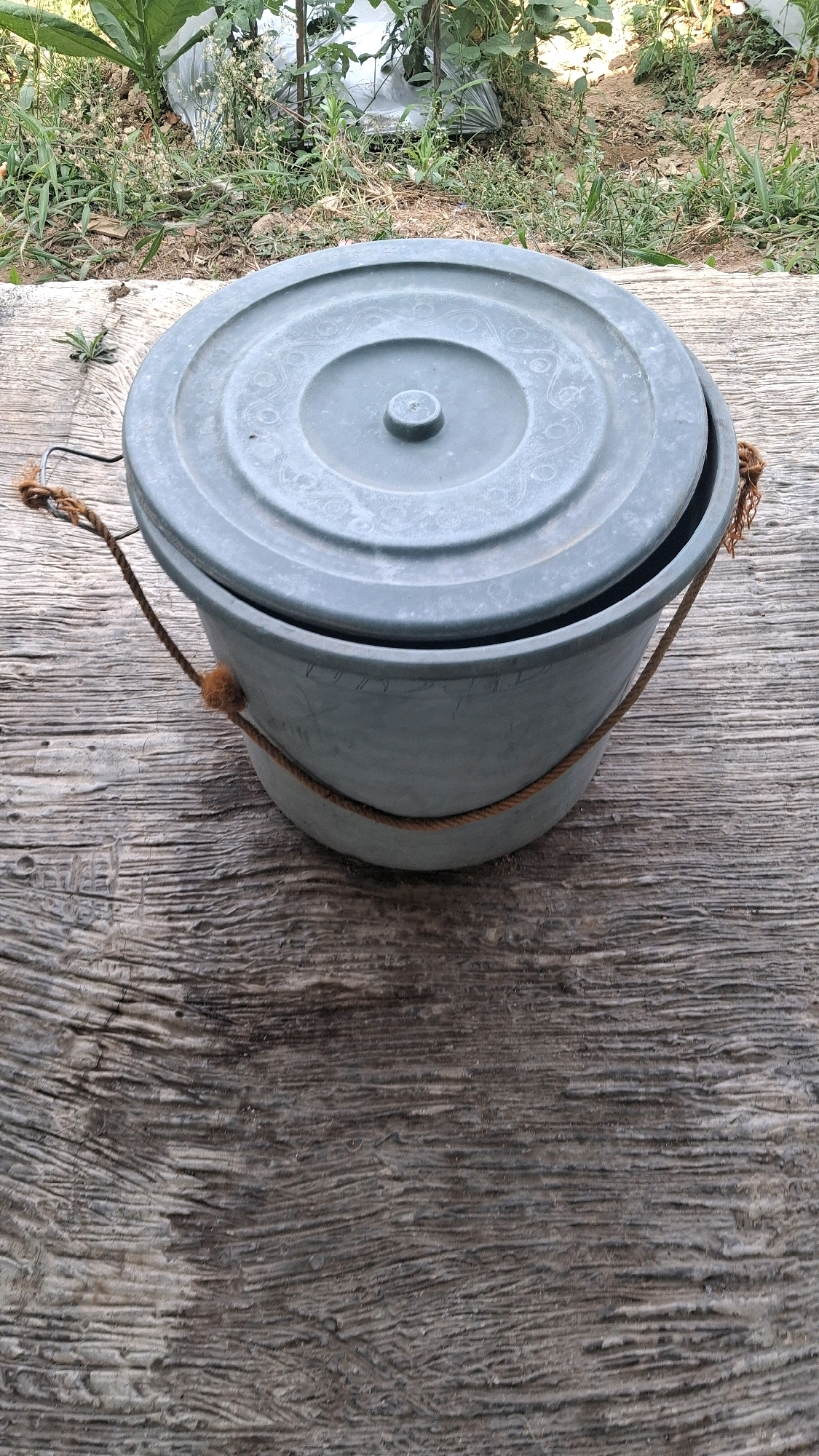 A grey plastic bucket with a lid sits on a textured wooden surface. A brown rope is tied around the bucket, with a knot on each side. The lid is slightly ajar. In the background, green plants and dry earth are visible. The scene is outdoors, suggesting a natural or semi-outdoor environment. The overall impression is of a simple, utilitarian object placed in a rustic setting. The specific location in Giriwarno, Indonesia is not discernable from the image itself.