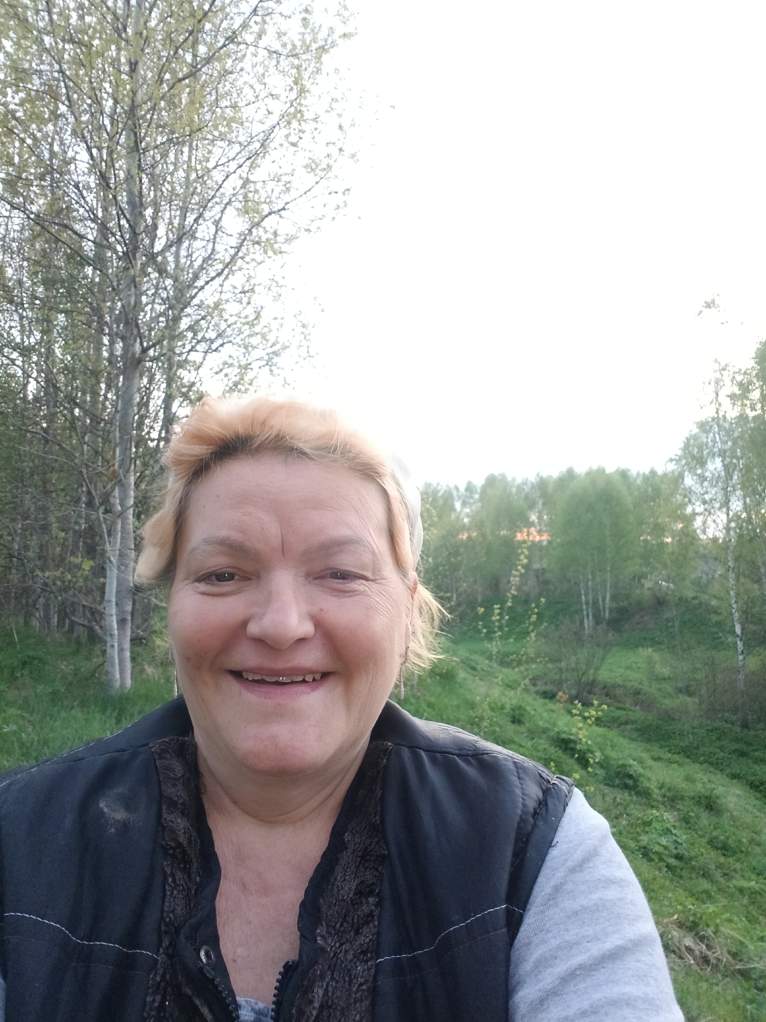 A woman is photographed outdoors. She has blonde hair and is wearing a dark jacket over a gray shirt. Her face is visible, and she is smiling. In the background, there are trees, suggesting a natural environment. The location is Красный Маяк, Russia.