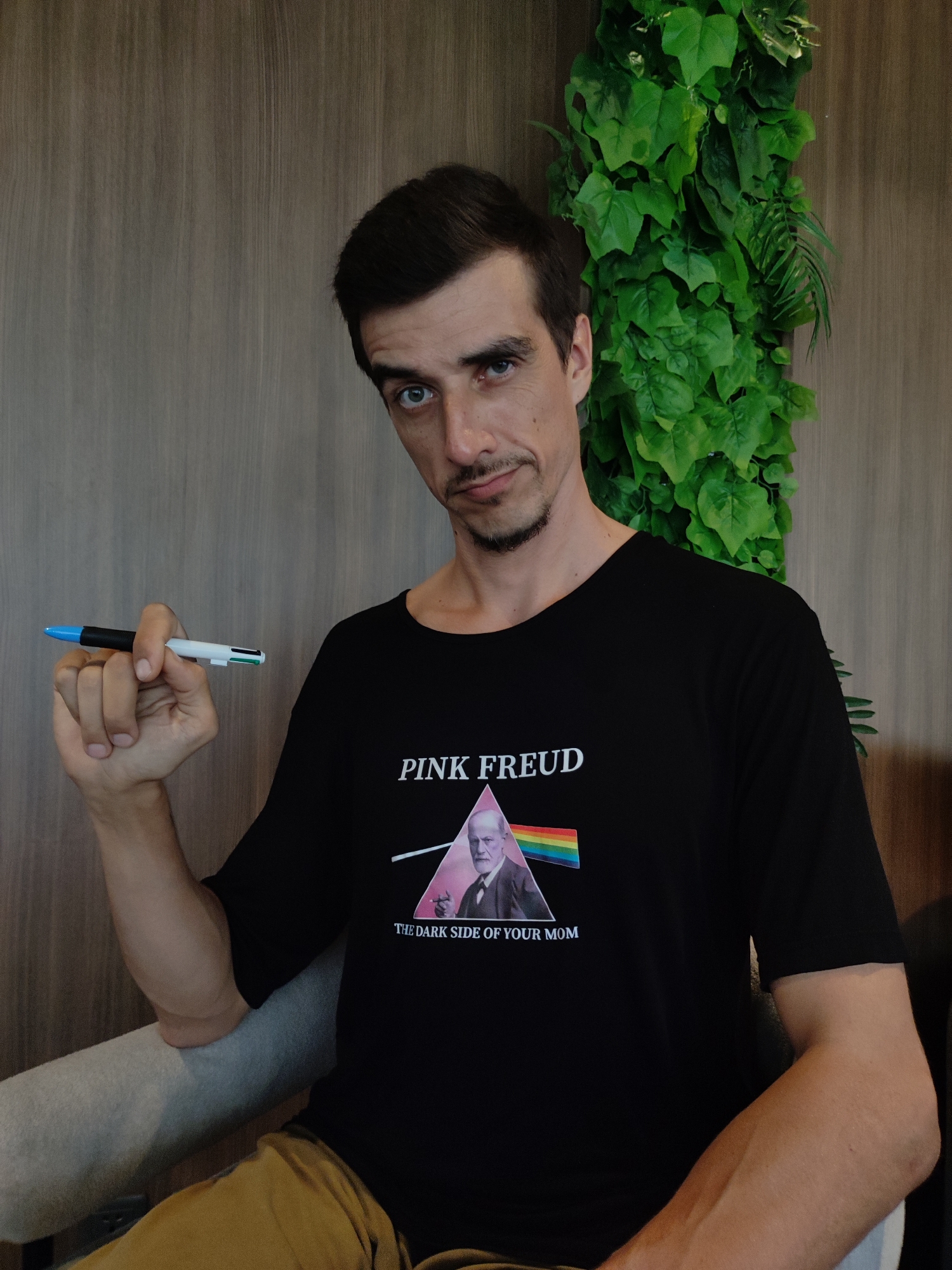 A man with dark hair, a mustache, and a goatee is seated indoors, likely in North Kuta, Indonesia. He looks directly at the camera with a subtle, intriguing expression. He wears a black t-shirt featuring a graphic that reads "PINK FREUD" above an image of Sigmund Freud within a prism, and "THE DARK SIDE OF YOUR MOM" below. In his right hand, he holds a multi-color pen with blue, black, white, green, and red elements. The background consists of vertical wooden wall paneling on the left, and a vibrant green artificial plant wall on the right. He is sitting in a gray armchair, with hints of brown trousers visible. The lighting suggests it is daytime.