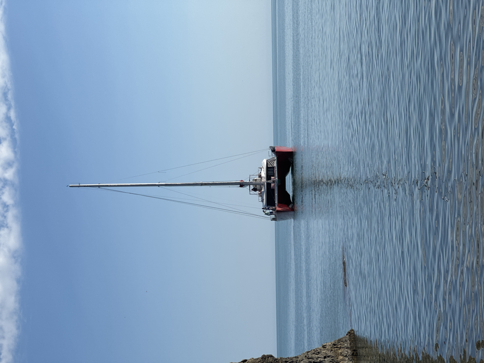 The image, when viewed upright, displays a tranquil coastal scene featuring a catamaran on calm, blue waters under a bright sky. A white and red catamaran, equipped with a tall mast and visible rigging, is positioned centrally, facing towards the right side of the frame. A single, indistinct person can be seen standing on the deck of the catamaran, near the stern.
The sky is a clear, light blue, with a strip of soft white clouds visible along the top left edge. The water is placid, exhibiting gentle ripples that reflect the serene sky. In the foreground, dark, rocky formations emerge from or lie beneath the water, suggesting a rugged coastline or seabed.
The scene depicts a peaceful moment, likely during late morning or early afternoon on a sunny day, with mild weather conditions. The catamaran appears to be at anchor or moored, and no active events are taking place. The setting aligns with a coastal location such as Divnomorskoe, Russia. There is no visible text in the image.