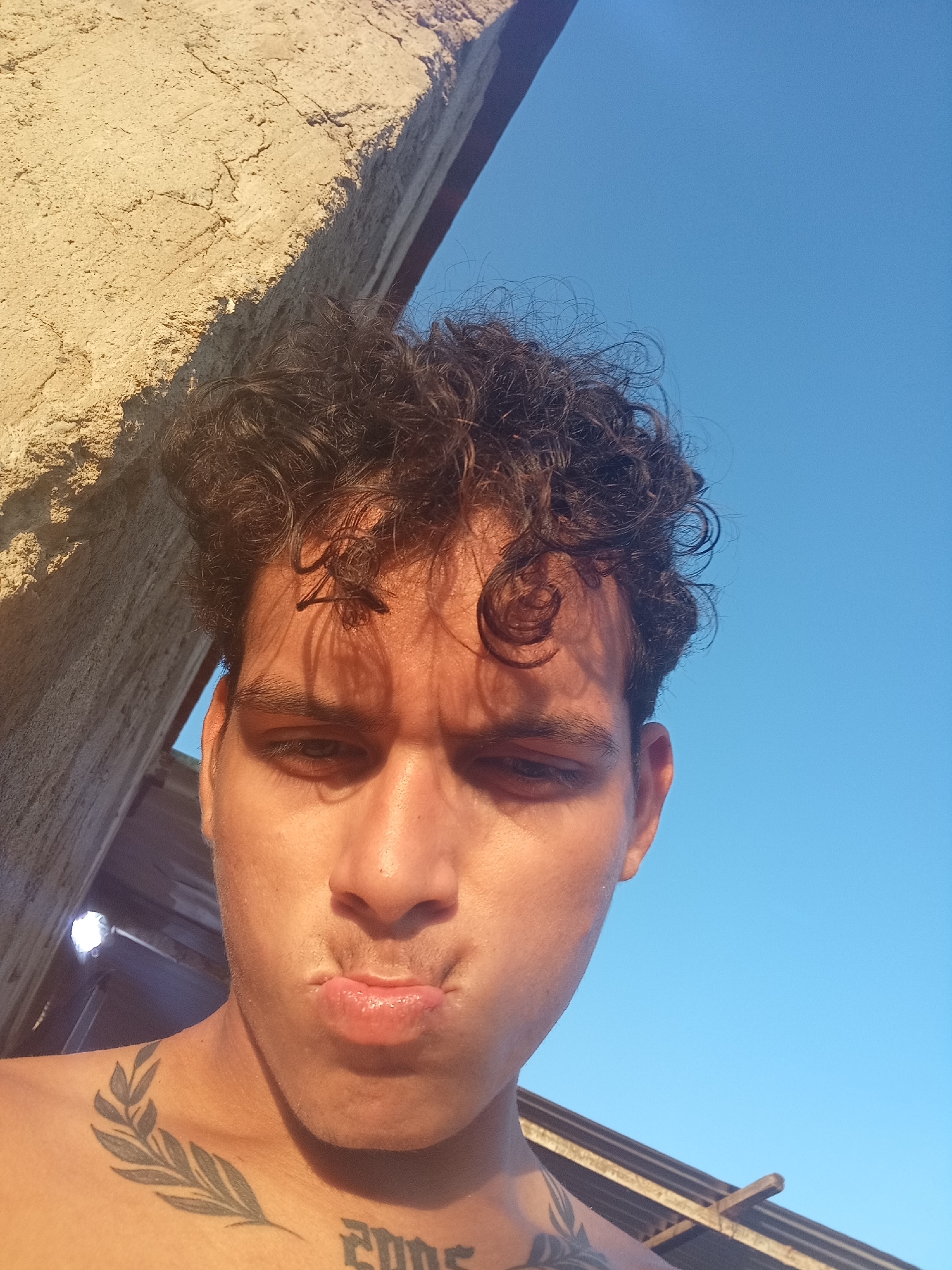A low-angle, medium shot depicts a young, shirtless man with dark, curly hair looking down at the camera with a pursed-lip expression. He appears to be outdoors. A stone-like structure is behind and to his left, while a clear, blue sky is visible to his right. The man has two tattoos: a leafy branch on his left shoulder and the word "2005" on his chest. The scene is bathed in strong sunlight. The location is presumed to be Altagracia, Venezuela, based on the prompt.