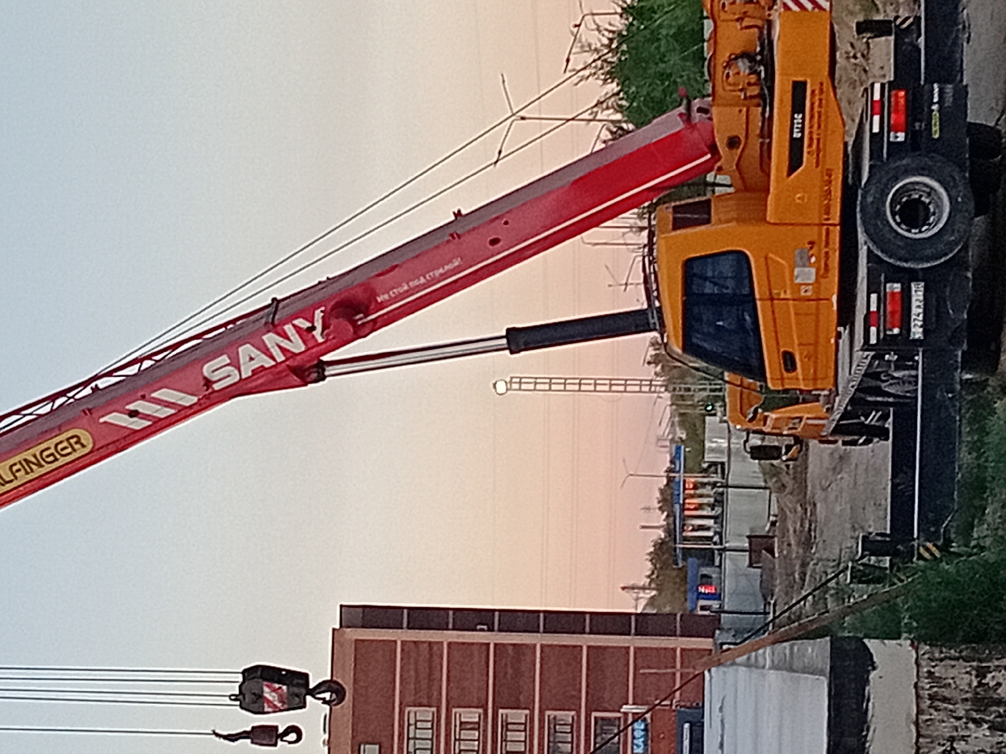 An outdoor, daytime, medium-shot image depicts a large red and yellow crane at a construction site. The crane's boom, labeled with the brand "SANY," extends diagonally across the frame from the upper right to the lower center. A yellow crane cabin is visible at the base of the boom, with a tire and a license plate that reads "T274 XR 16." To the left of the crane is a multi-story brick building with rows of windows. In the background, there are power lines, streetlights, and some industrial structures, suggesting an urban or industrial area. The sky is a soft, light orange and blue, indicating either dawn or dusk. The ground appears to be unpaved, with some dirt and grass. The overall scene suggests ongoing construction activity in the city of Semey, Kazakhstan.