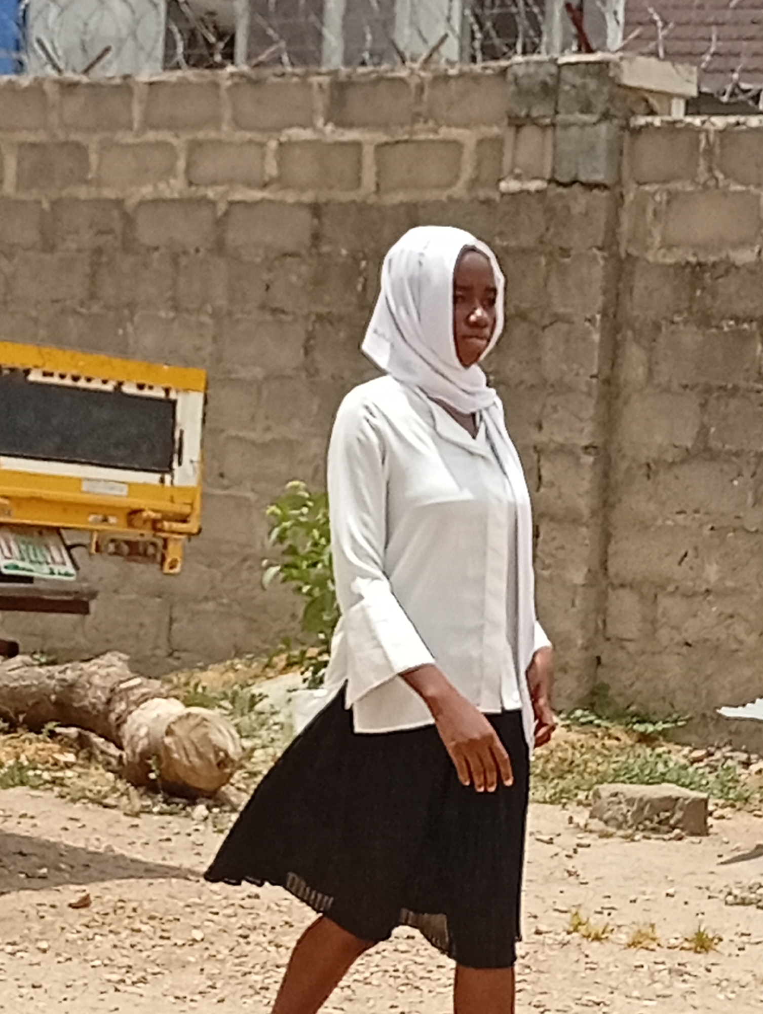 A young woman, likely in her late teens or early twenties, is walking in an outdoor location in Lokoja, Nigeria. She is wearing a white headscarf, a white button-down shirt, and a black pleated skirt. The environment suggests an urban or peri-urban setting. In the background, there is a cinder block wall topped with what appears to be barbed wire. To the left of the woman, there is part of a yellow vehicle and a log on the ground. The ground is sandy and appears dry. The time of day seems to be bright daylight.