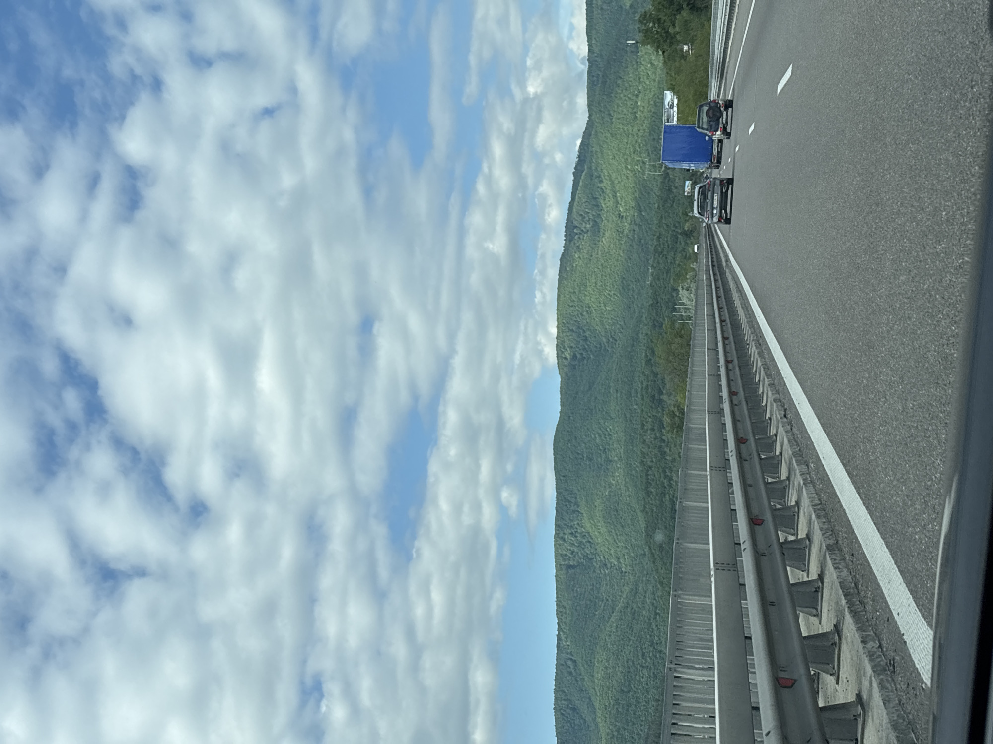 The image shows a view from a car traveling on a highway, possibly a bridge or elevated section, near Gorno Varshilo, Bulgaria. The landscape includes a mountain range covered in green vegetation and a sky filled with scattered white clouds against a blue background. On the right side of the image is a section of the highway with a guardrail. Two vehicles, including a blue truck, are visible on the road. The scene appears to be daytime, with natural light.