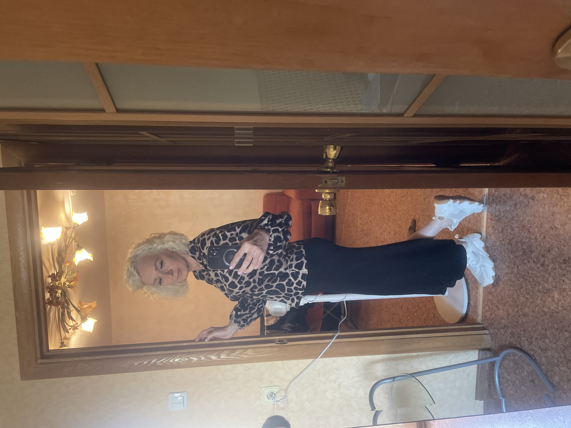 A woman with short, blonde, curly hair is standing in a doorway, taking a mirror selfie with her smartphone. She is wearing a leopard-print long-sleeved shirt and a long black skirt. She is also wearing white sneakers with thick soles. The background appears to be an apartment or house, with a wooden door frame and a chandelier visible in the reflection of the mirror. A floor fan is positioned behind her. The floor is covered in a cork material. The image is taken indoors.