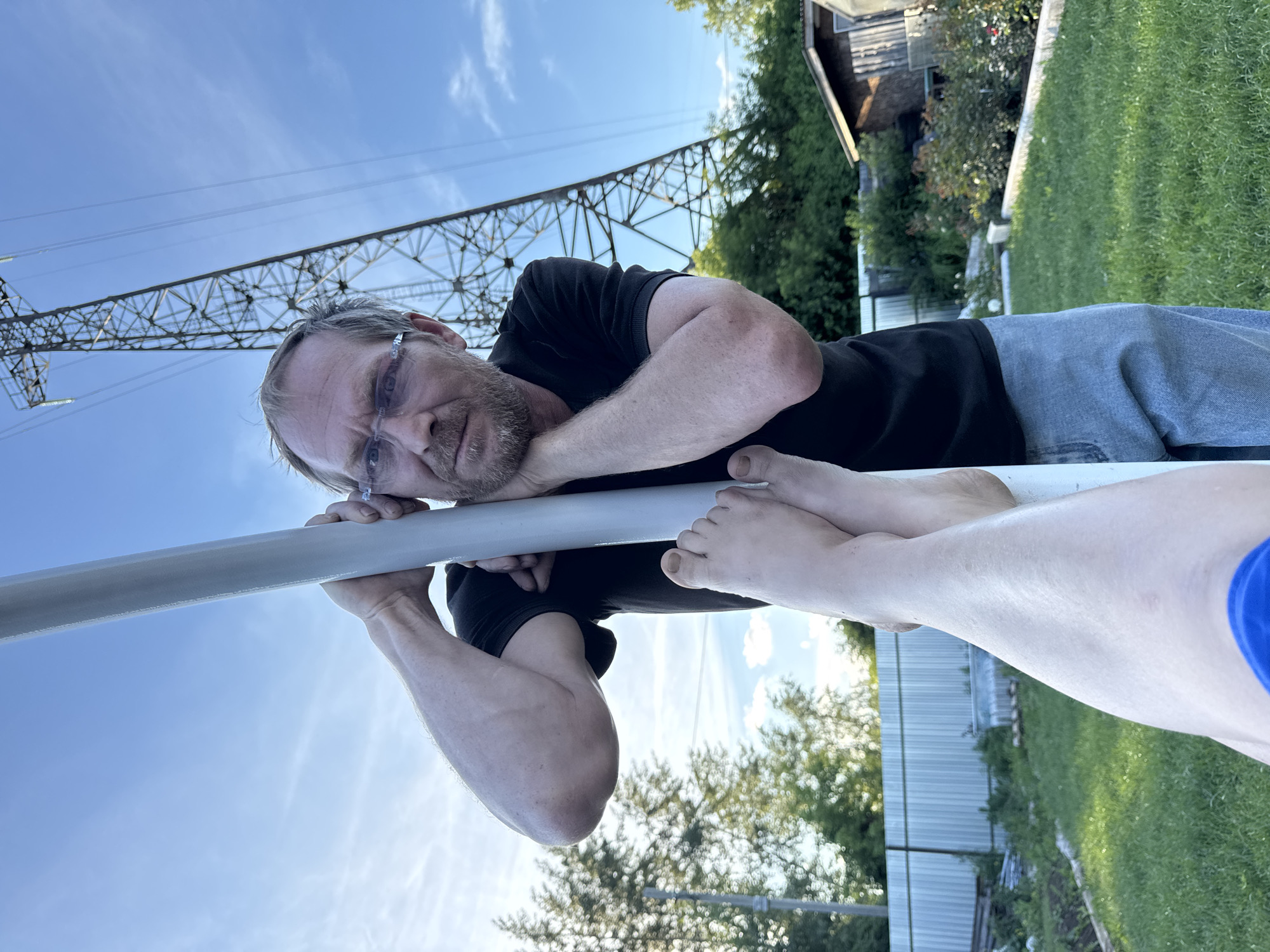 In Chaikovsky, Russia, a man with glasses and a beard leans against a white pole in a yard. He is wearing a black t-shirt and blue jeans. His arms are resting on the pole, and his head is tilted, looking towards the camera. In the foreground, a pair of bare feet are visible, one leg extended towards the camera. Behind the man, a large metal electrical tower dominates the sky, with trees and a small wooden shed in the background. The sky is blue with some wispy clouds. The ground is covered with green grass.