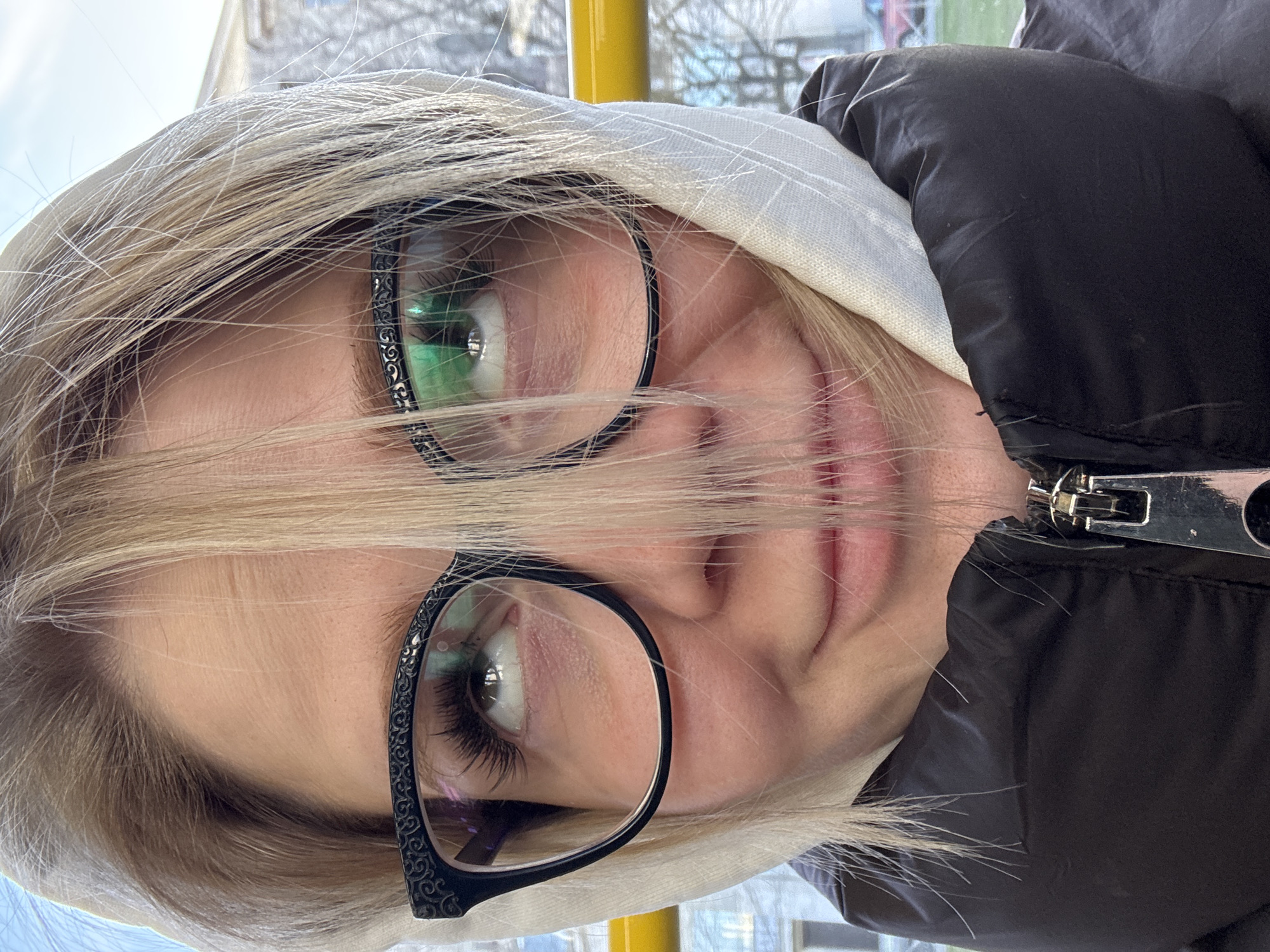 A close-up, slightly rotated image of a woman wearing glasses and a hooded jacket. The woman has blonde hair and light skin. She is wearing black framed glasses. The hood is light gray or white. She is wearing a black puffy jacket with a metal zipper pull visible. The background is blurred, but there is evidence of an exterior scene through a window. Based on the visible yellow pole and the blurry background, it's possible she is on public transport. Considering the location as Saint Petersburg, Russia, this is a likely scenario.