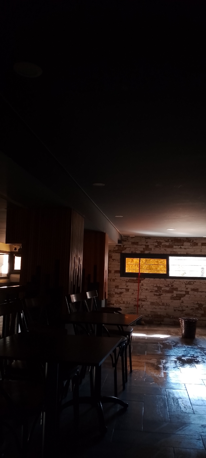 A dimly lit interior space reveals a section of tables and chairs in the foreground, shrouded in shadow. The far end of the room is illuminated by sunlight streaming through windows. A brick wall features two windows, one casting a warm, yellow light due to a screen. The floor appears wet or reflective, adding brightness to the area near the windows. A broom stands near one window, and a bucket rests on the floor.