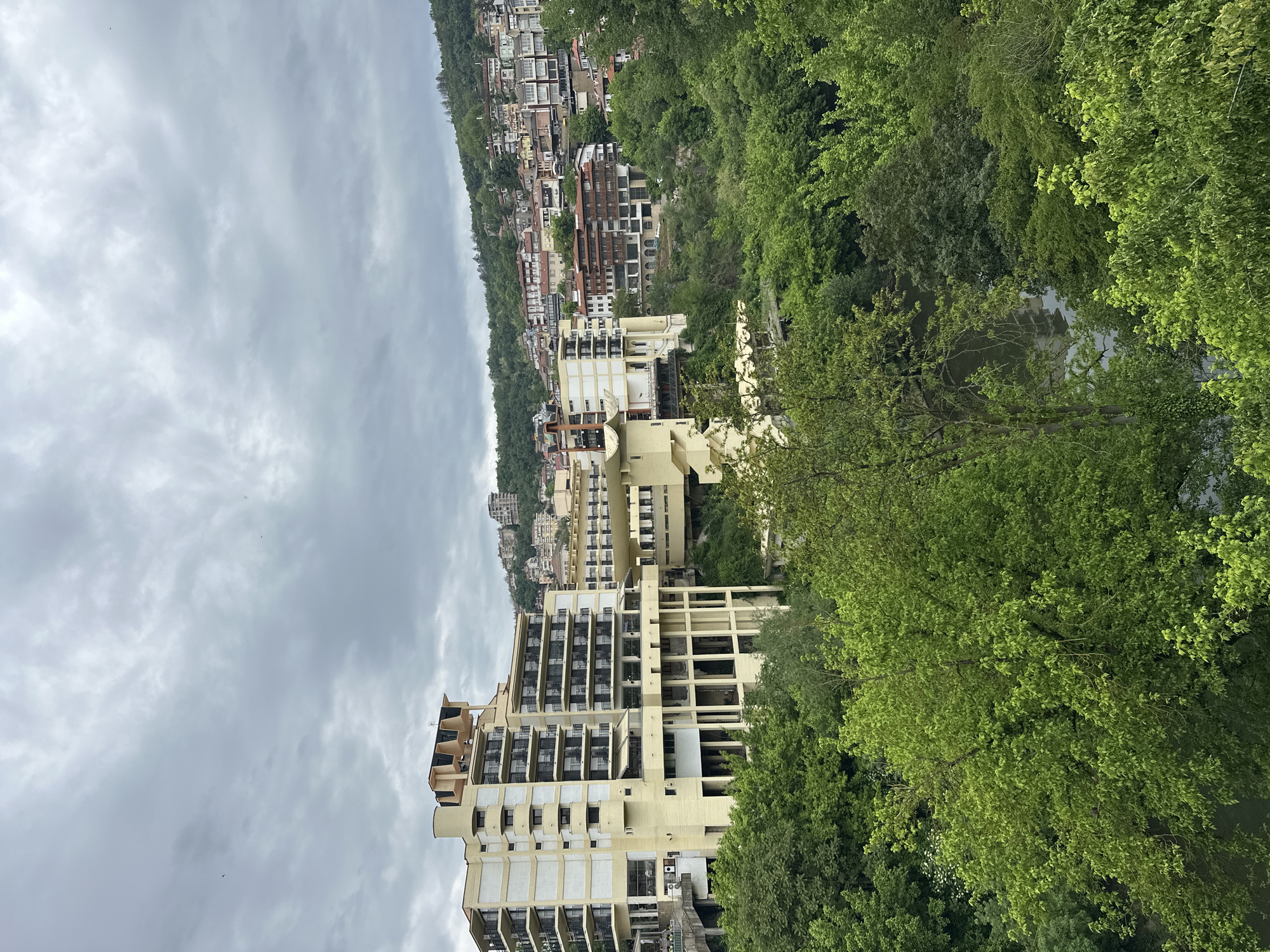 The image shows a high-angle view of Veliko Tarnovo, Bulgaria, featuring a light-colored, multi-story hotel building nestled among green trees on a hillside. A river or stream is visible through the trees to the right. In the background, a densely populated hillside is filled with various buildings. The sky is overcast.