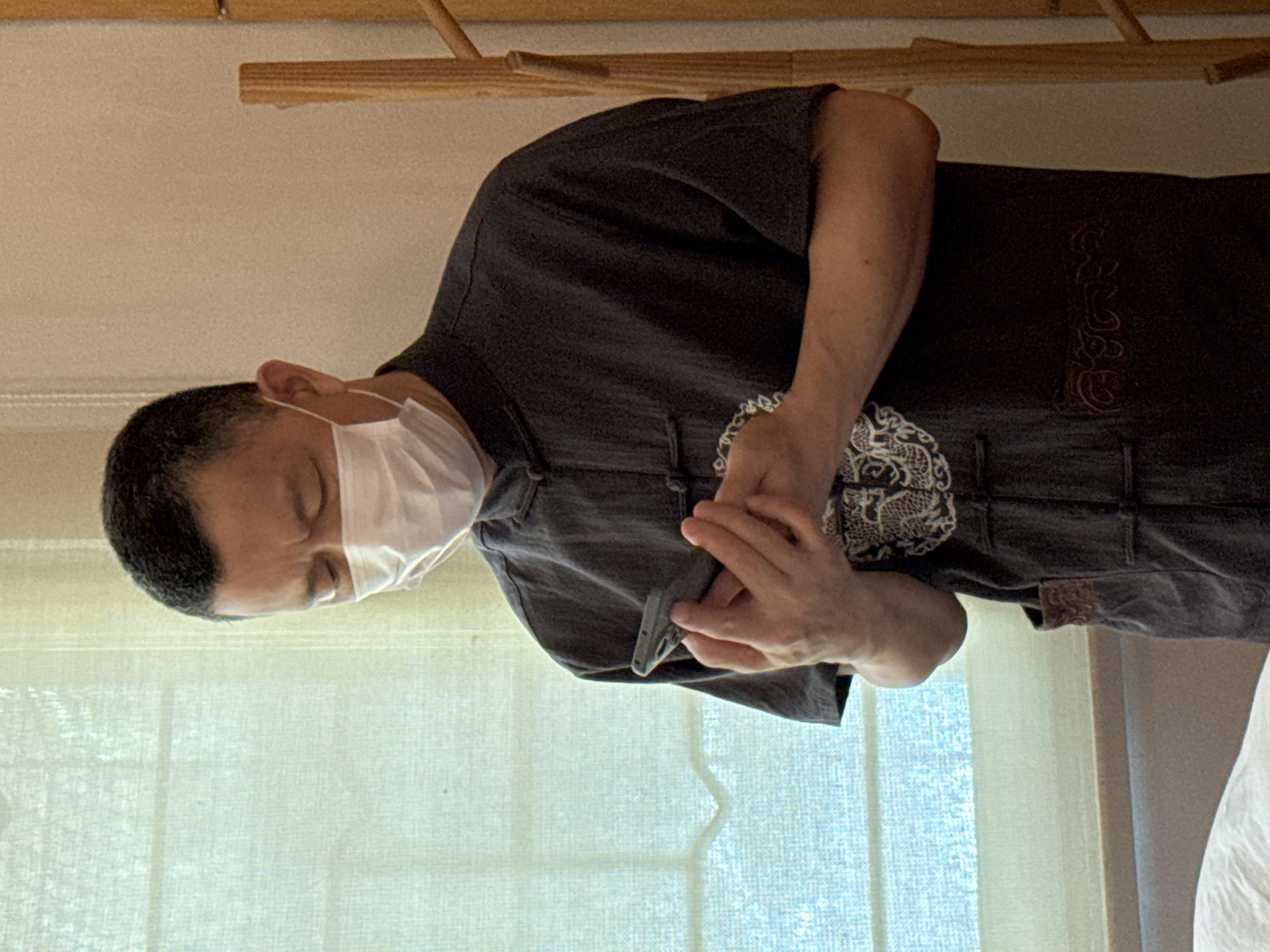 A man wearing a dark grey, traditional Chinese-style shirt and a white medical mask is standing indoors, looking down at a smartphone he is holding in his hands. He has short dark hair and appears to be middle-aged. The background shows a sheer, light-colored curtain or window covering and a portion of a light-colored wall. To the right, a vertical wooden structure with horizontal pegs is visible. The overall lighting suggests an indoor environment, possibly a treatment room or a spa, given the presence of what appears to be a bed or massage table in the lower left foreground. The man is focused on his phone, with his brow furrowed.