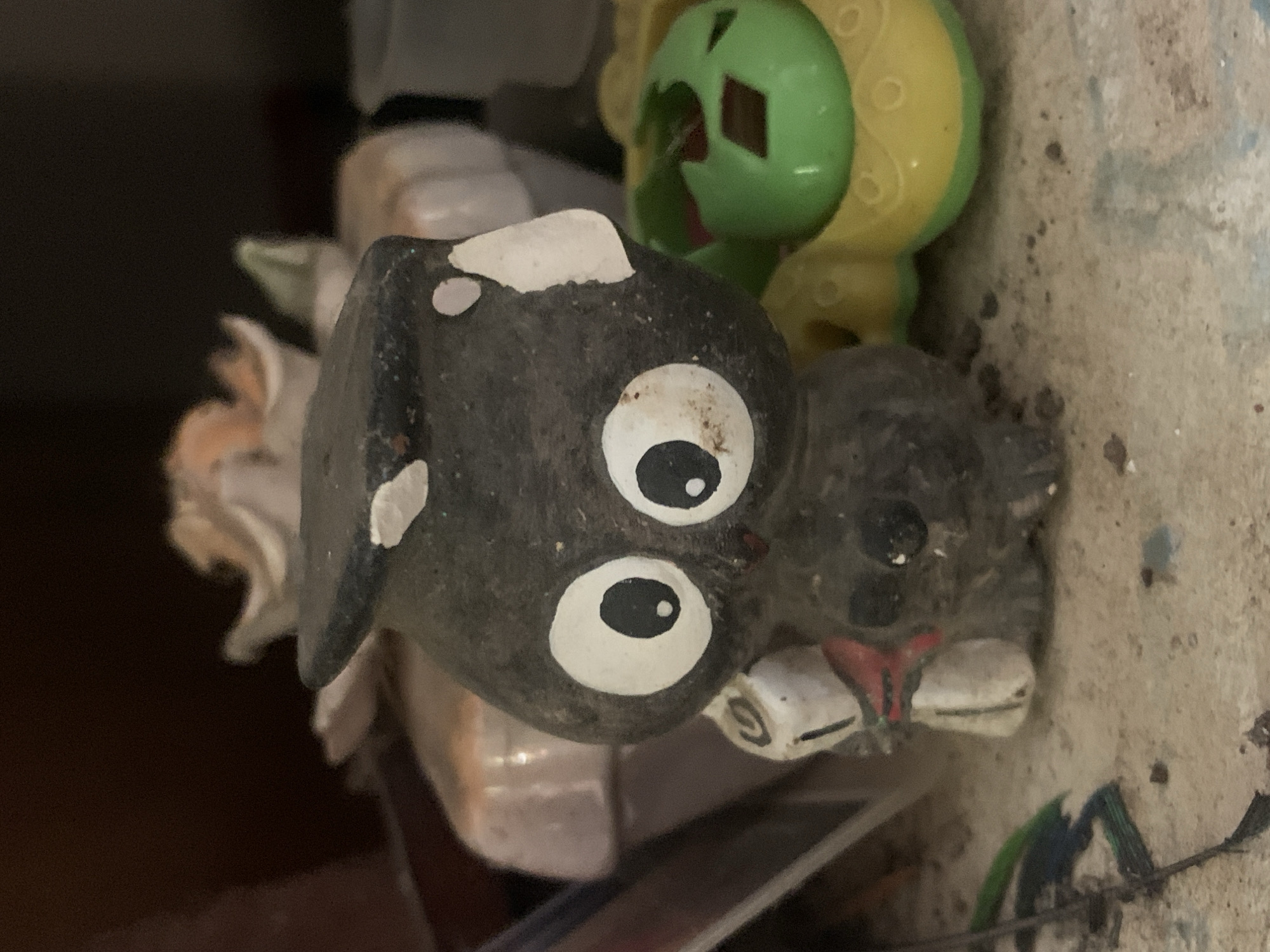 The image shows a close-up of several objects. The most prominent is a small, possibly ceramic or stone, figurine of a black and white dog with large, round eyes. The dog has white markings on its head and face. To the upper right of the dog figurine, there is a green and yellow plastic toy. The dog appears to be resting on or near a surface that is cluttered with other items, including what looks like a faded flower arrangement or decoration to the left and some shiny or reflective materials underneath. The background is mostly out of focus and indistinct, with a dark area to the left. There is no indication as to the location of the image, and thus the city of Santa Ana, Venezuela cannot be substantiated from the image.