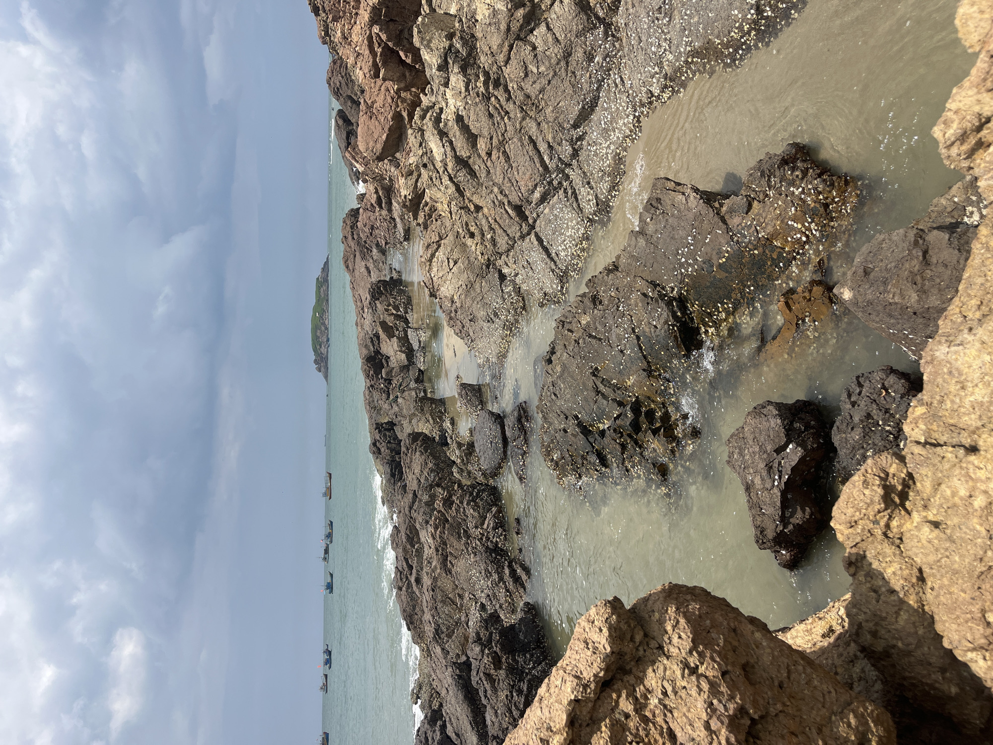 The image shows a rocky coastline with calm water and several small boats in the distance. The foreground is dominated by large, rough, reddish-brown rocks, some of which are partially submerged in shallow, murky water. The rocks are textured and appear to be covered in small white shells in some areas. The water is a light greenish-brown and reflects the overcast sky. In the mid-ground, the water recedes to reveal more rocks and sandy patches. Further out, the ocean is a muted teal color, with several small fishing boats scattered across the surface. The sky is cloudy and grey, suggesting an overcast day. In the far distance, a small, green, tree-covered island with some buildings is visible on the horizon. This scene is characteristic of the Mui Ne coastline in Vietnam, known for its dramatic rock formations and fishing villages. There are no people visible in the image.