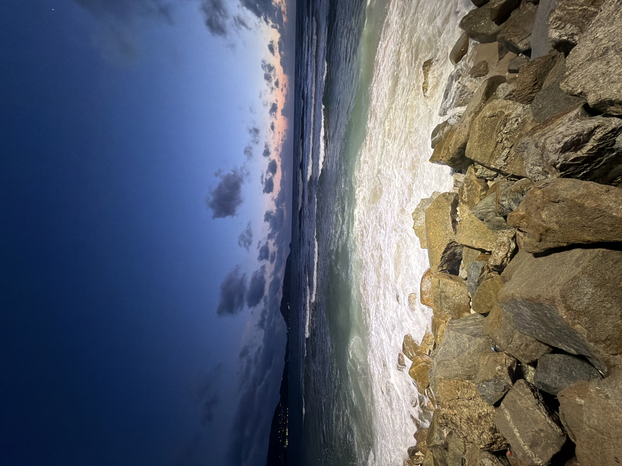This is a nighttime, low-angle, long-exposure photograph of a rocky shoreline in Patong, Thailand. The foreground is dominated by large, rough-hewn rocks and boulders, some of which are wet from the encroaching waves. The ocean is in the midground, with white, foamy waves crashing against the shore and receding over the rocks. The water is a deep blue-green, with lighter turquoise hues where the foam is present. In the background, the dark silhouette of a coastline with distant lights can be seen, suggesting the presence of the city of Patong. The sky above is a deep twilight blue, transitioning to lighter shades of pink and orange near the horizon, with scattered dark clouds. The overall scene evokes a sense of natural beauty and the powerful presence of the ocean at night.