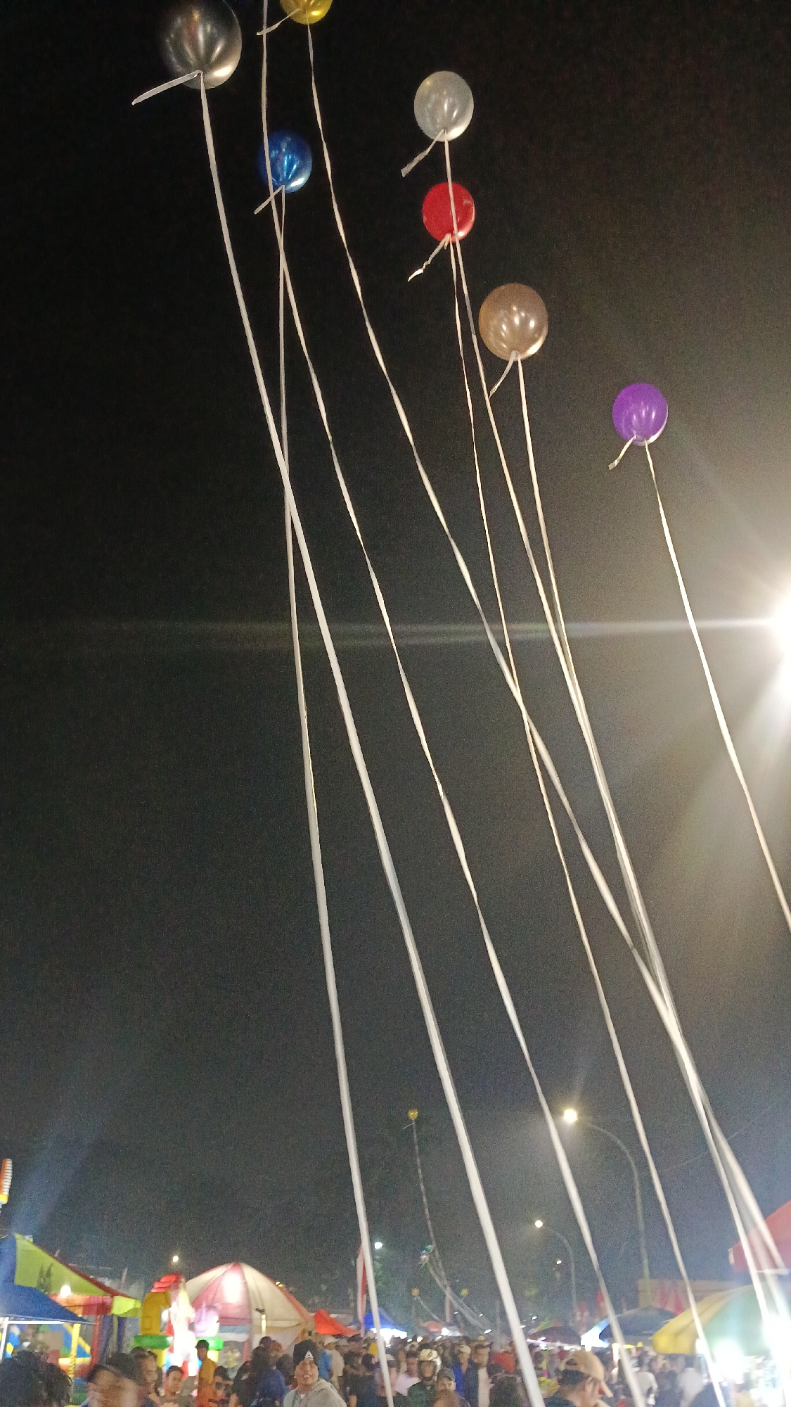 In Tajurhalang, Indonesia, a nighttime outdoor event is underway, possibly a festival or fair. A dark sky serves as the backdrop for several colorful balloons, each trailing a long white ribbon. The balloons are silver, yellow, blue, clear, red, brown, and purple, some appearing slightly translucent.

Below the balloons, the scene is populated with numerous people gathered around stalls and tents, suggesting a marketplace or entertainment area. The lighting is dim, with some artificial illumination from streetlights and the bright glow of a light source to the right, possibly a large lamp or spotlight. The presence of tents with colorful canopies indicates various vendors or activity booths. The overall atmosphere appears to be lively, with many individuals milling about.