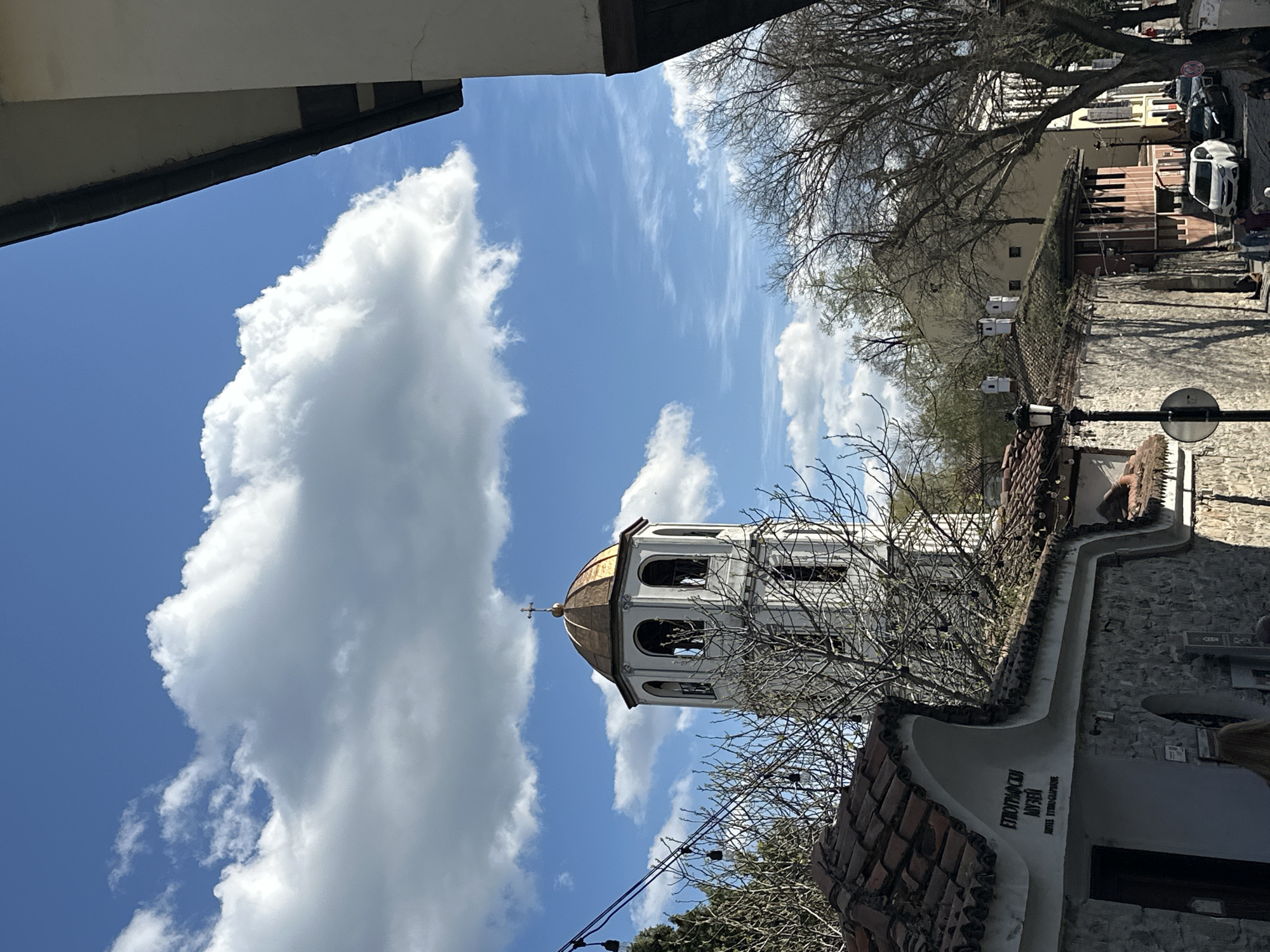 The image captures a low-angle view of the Ethnographic Museum in Plovdiv, Bulgaria, prominently featuring the museum's distinctive bell tower. The tower, painted white with a golden dome and a cross at the top, is set against a backdrop of a vibrant blue sky filled with large, fluffy white clouds. A portion of the building's stone and tile roof are visible. A bare tree is partially obscuring the tower. Adjacent to the building, other buildings and trees, also bare, line the street. The background reveals glimpses of buildings, parked cars, and pedestrians on the street. The photo is framed by a building on the left and some trees on the right.